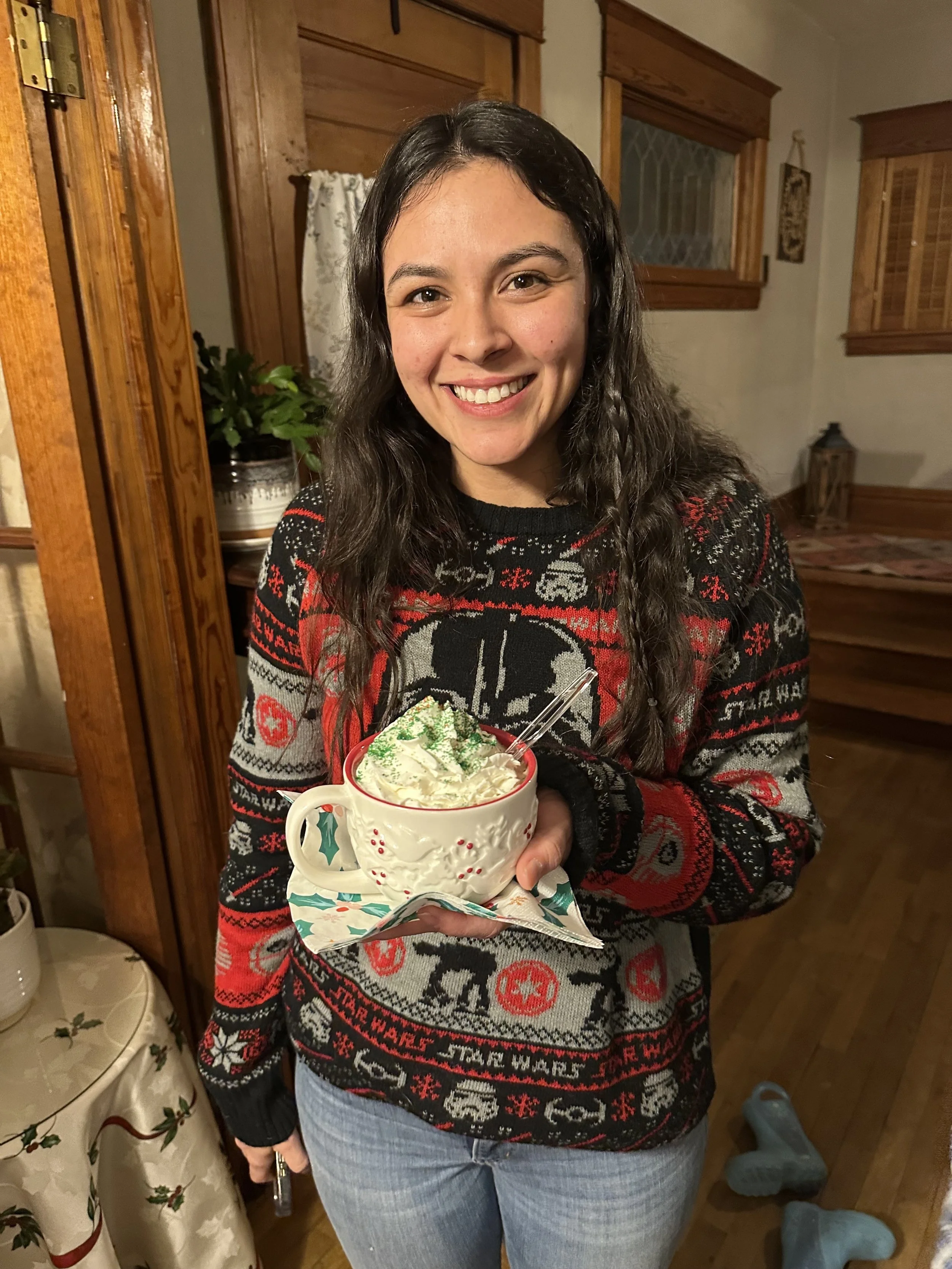 Young woman smiling, holding a large mug of whipped cream-topped hot chocolate with green sprinkles, in a cozy, wood-paneled room decorated for Christmas.