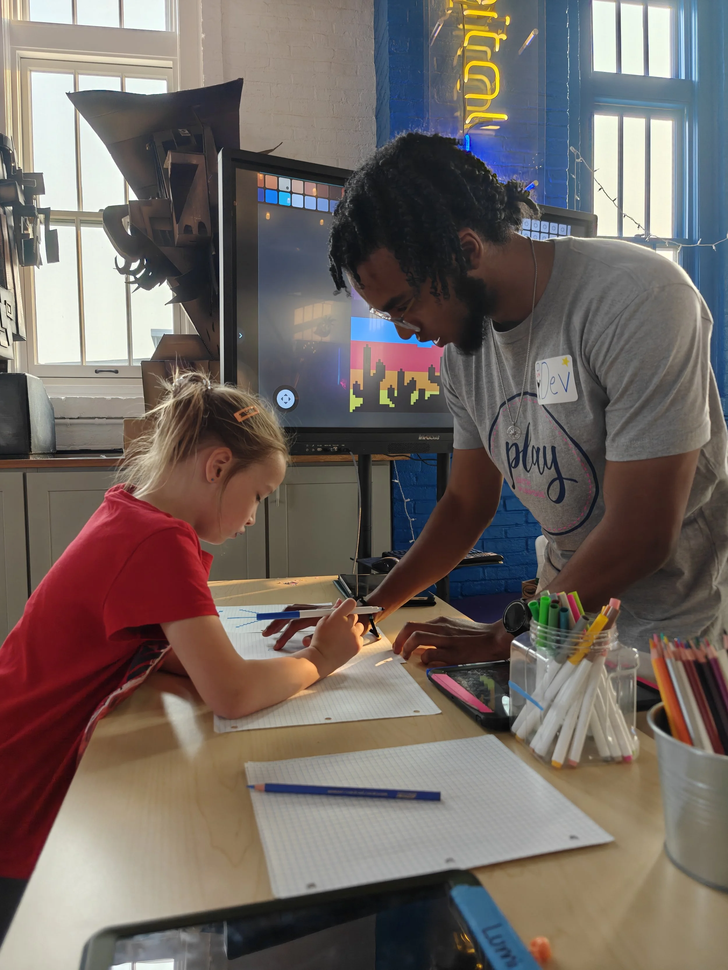 A young girl drawing on a notebook at a table while an adult male assists her, with various colored pens and paper in front of them, in a brightly lit room with blue walls and a large monitor in the background.