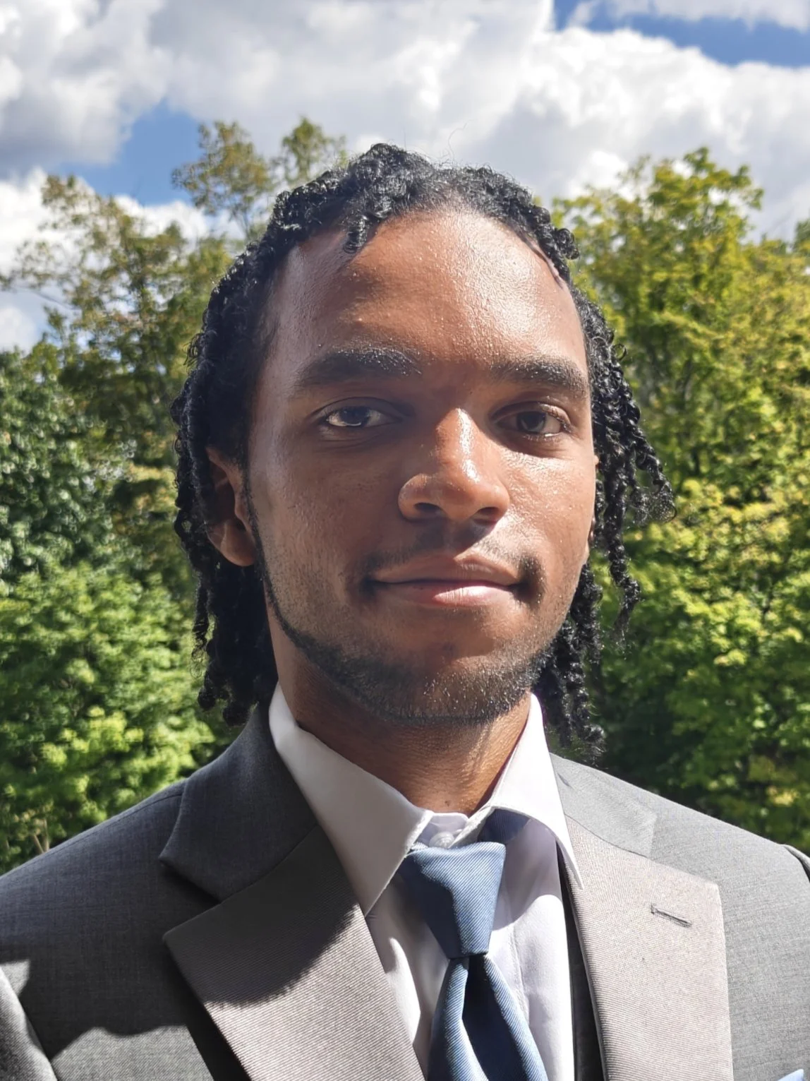 A young man wearing a grey suit, white shirt, and blue tie standing outdoors with green trees and a partly cloudy sky in the background.