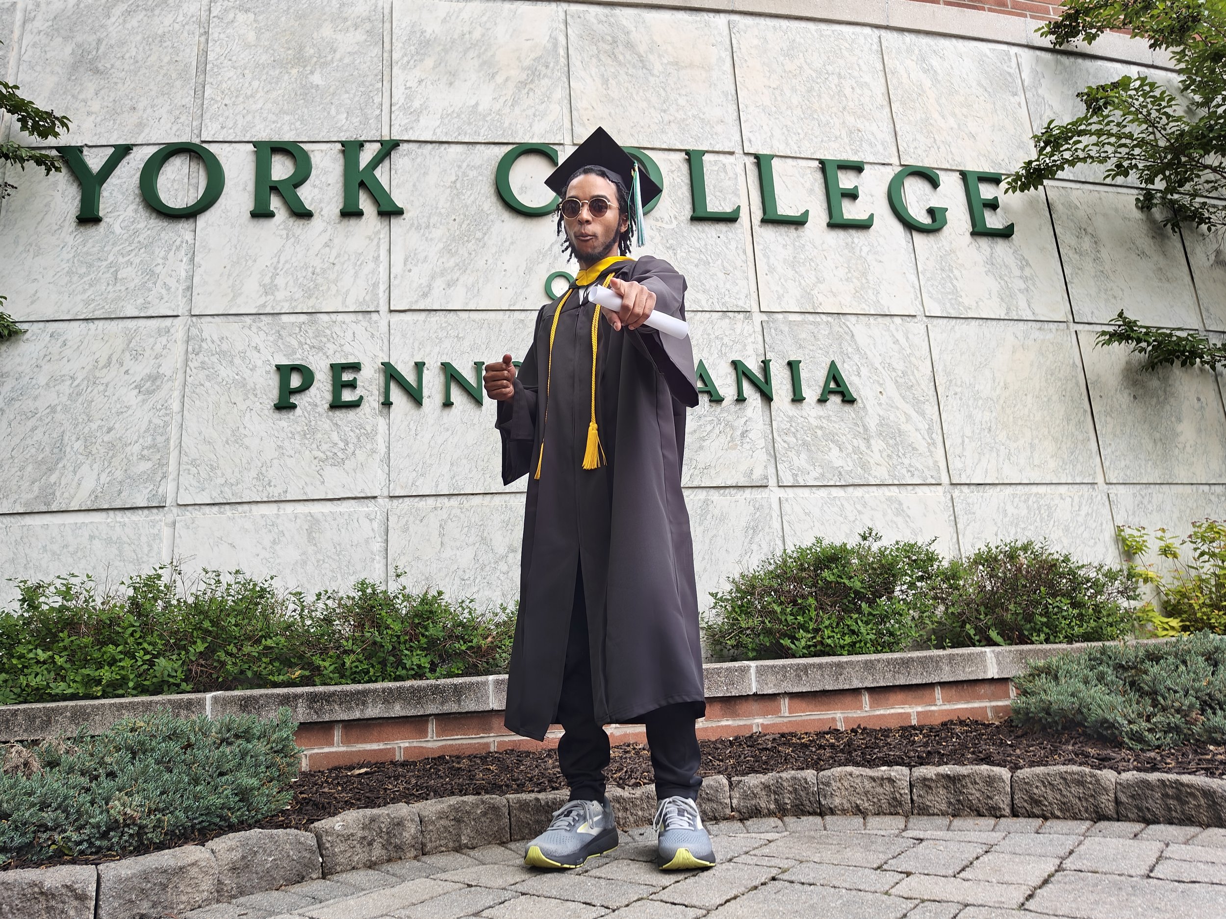 Graduate in cap and gown holding diploma pointing towards the camera in front of York College Pennsylvania sign.