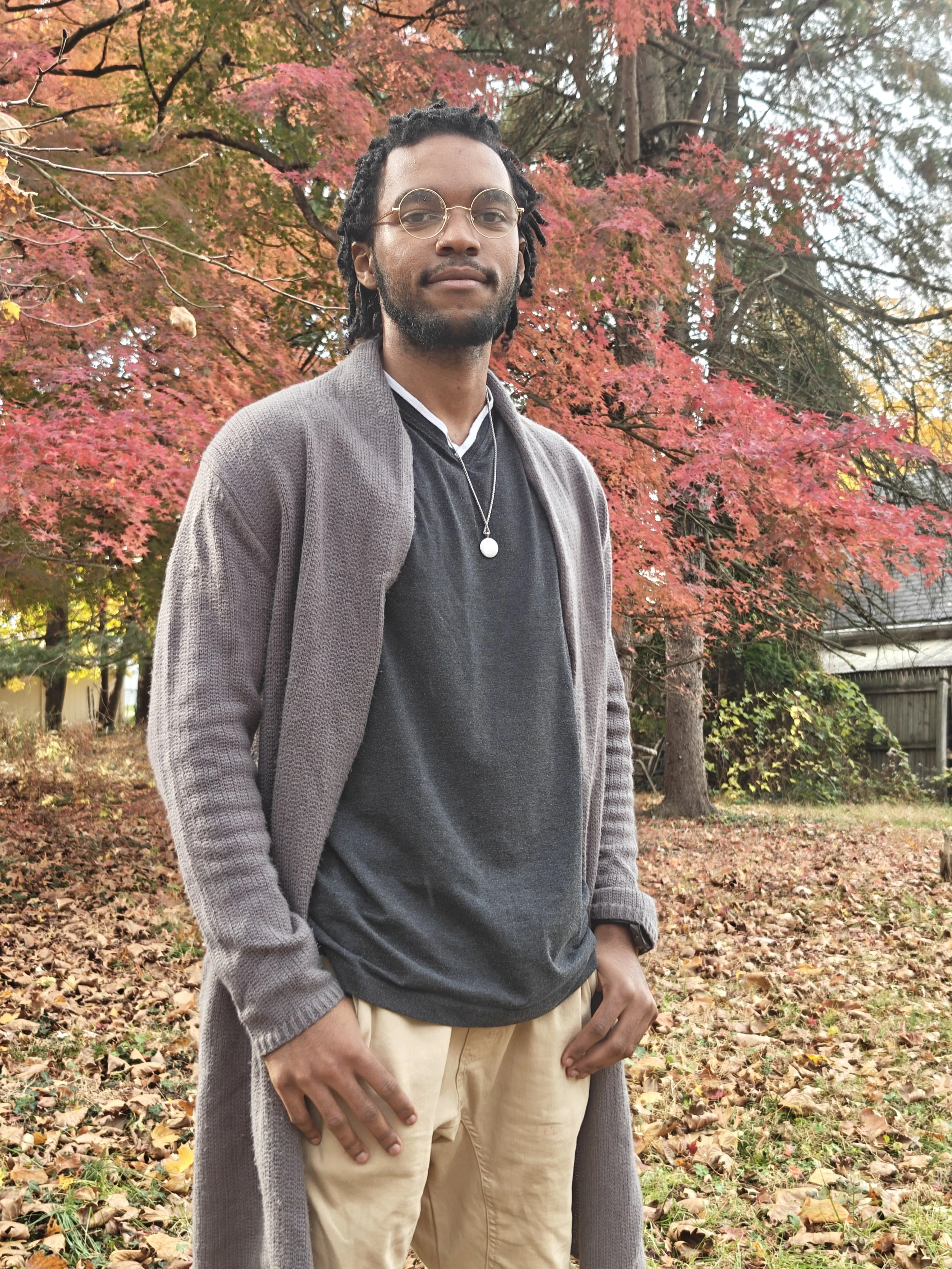 A man standing outdoors in front of trees with red and orange fall foliage, wearing glasses, a gray cardigan, a dark gray shirt, beige pants, and a pendant necklace.