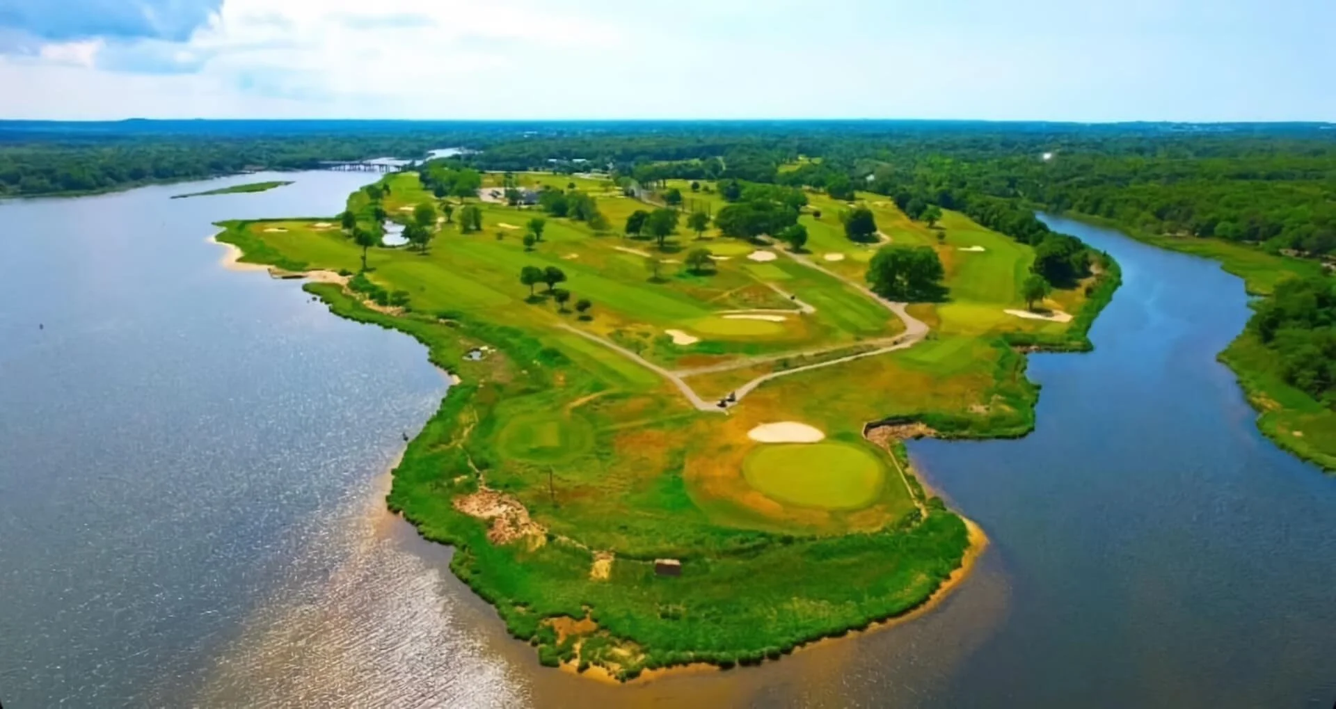 Aerial view of a golf course on a peninsula surrounded by water on three sides, with green fairways, sand bunkers, and scattered trees.