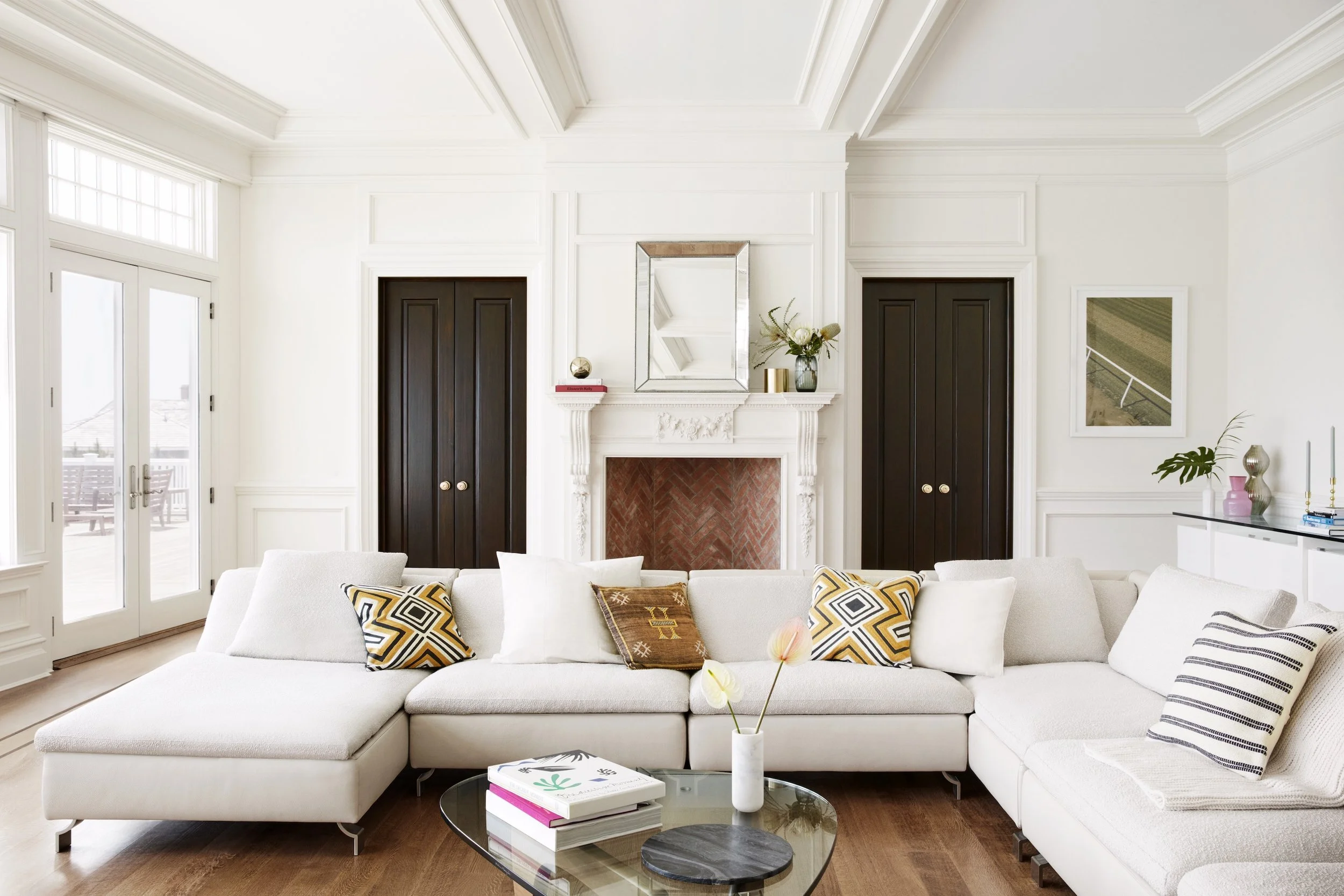 Modern living room with white walls, large beige sectional sofa with black, white, and yellow patterned pillows, and a round glass coffee table with books and a vase of tulips. There is a white fireplace with intricate molding and a mirror above it, flanked by two black doors. Large windows and glass doors let in natural light, and there is a white sideboard with decorative items and a framed picture on the wall.