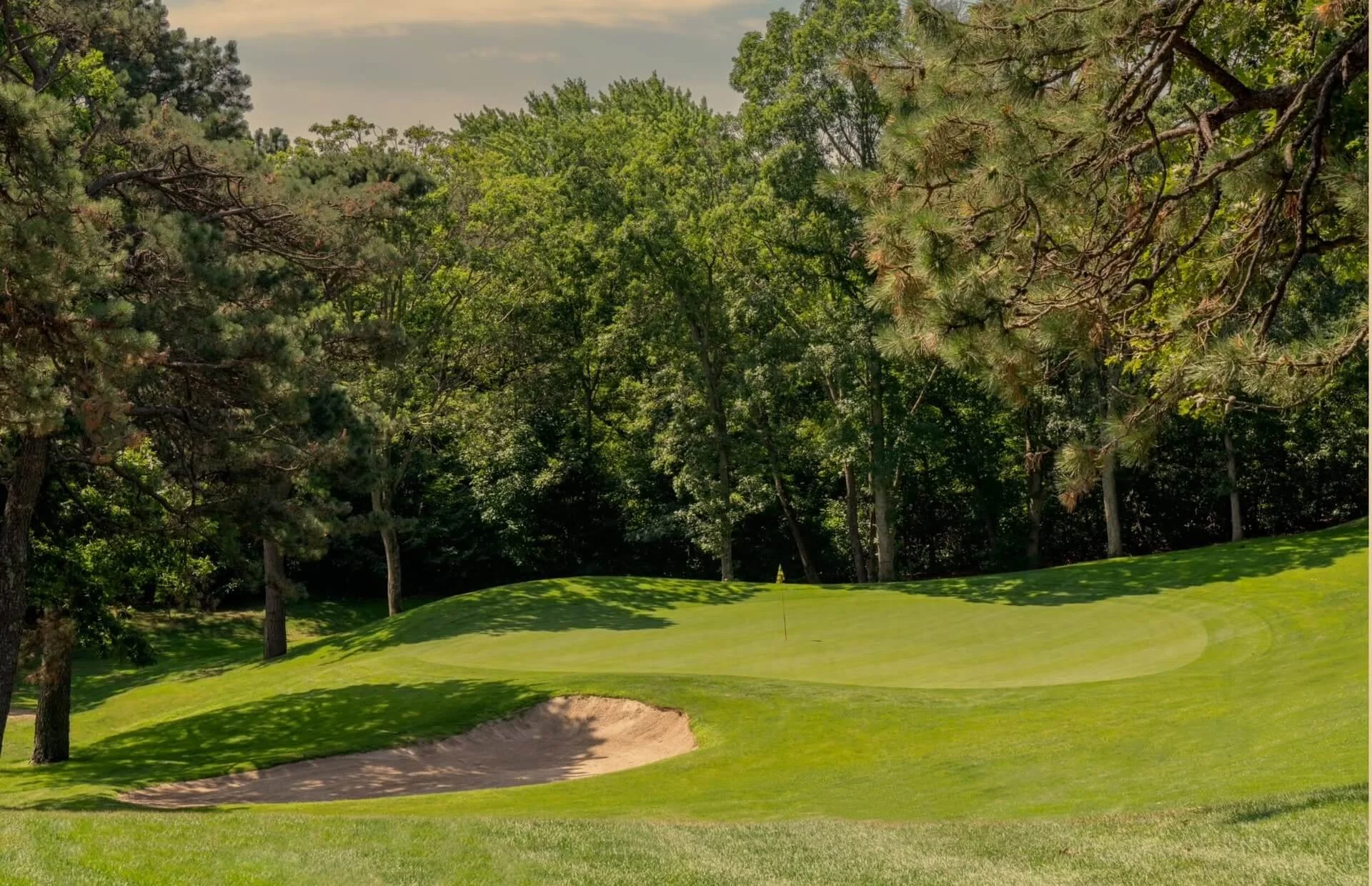 A golf course with a green and sand trap surrounded by trees under a partly cloudy sky.