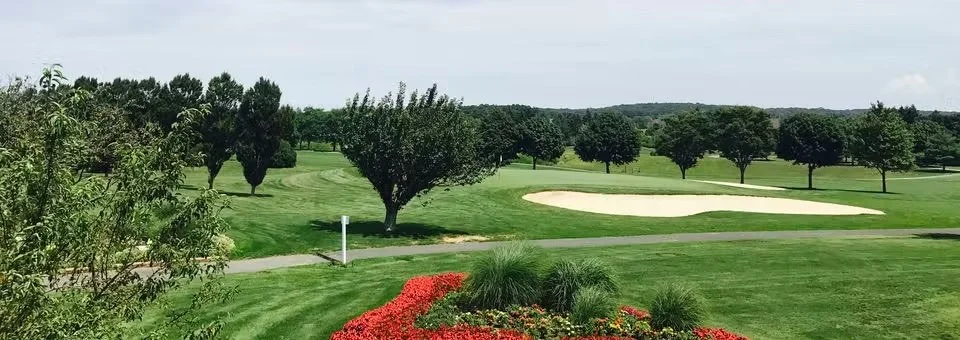 A golf course with neatly maintained green grass, trees, a sand bunker, a small path, and colorful flowers in the foreground.