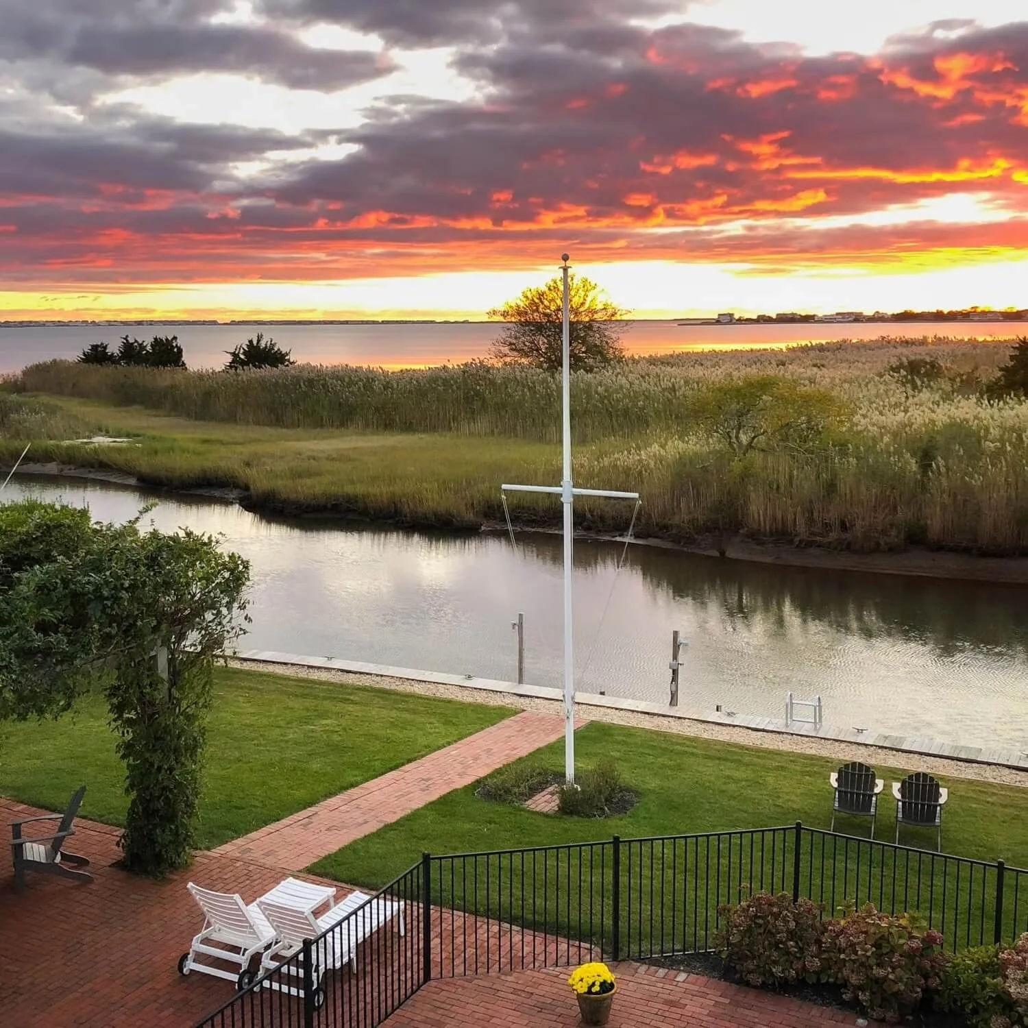Sunset over a river with a dock, grassy yard, and outdoor seating area, including Adirondack chairs and a flagpole.