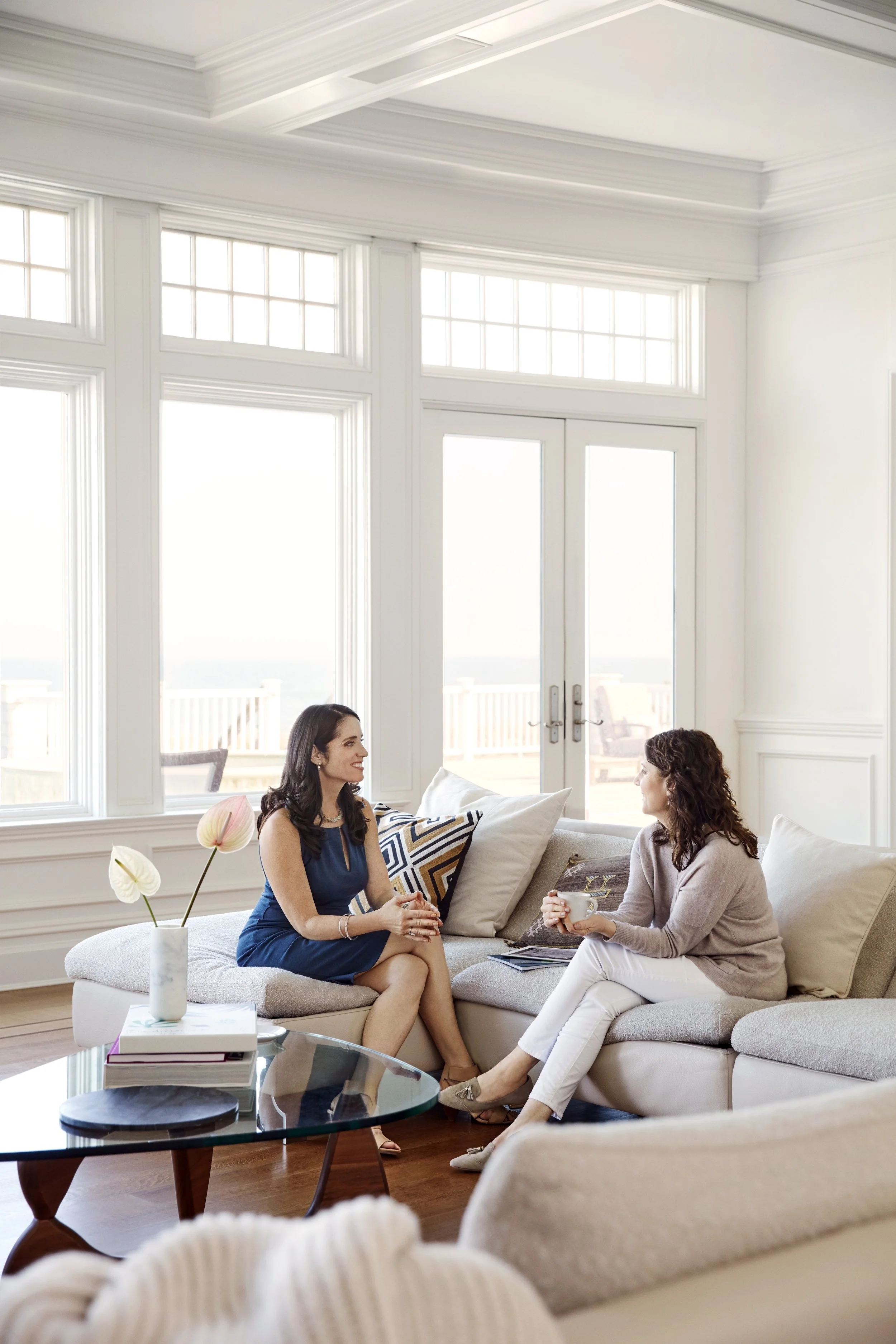 Two women sitting on a light-colored sofa, talking in a bright living room with large windows and a glass door, decorated with pillows, a flower vase, and books on a coffee table.
