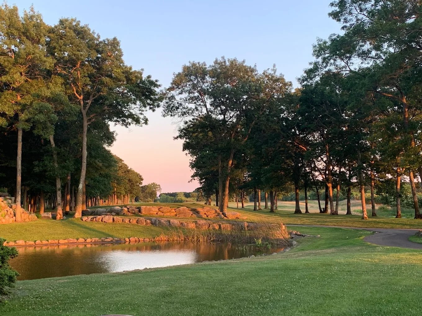 A scenic view of a golf course at sunset with green grass, a small water hazard, and tall trees along the fairway.