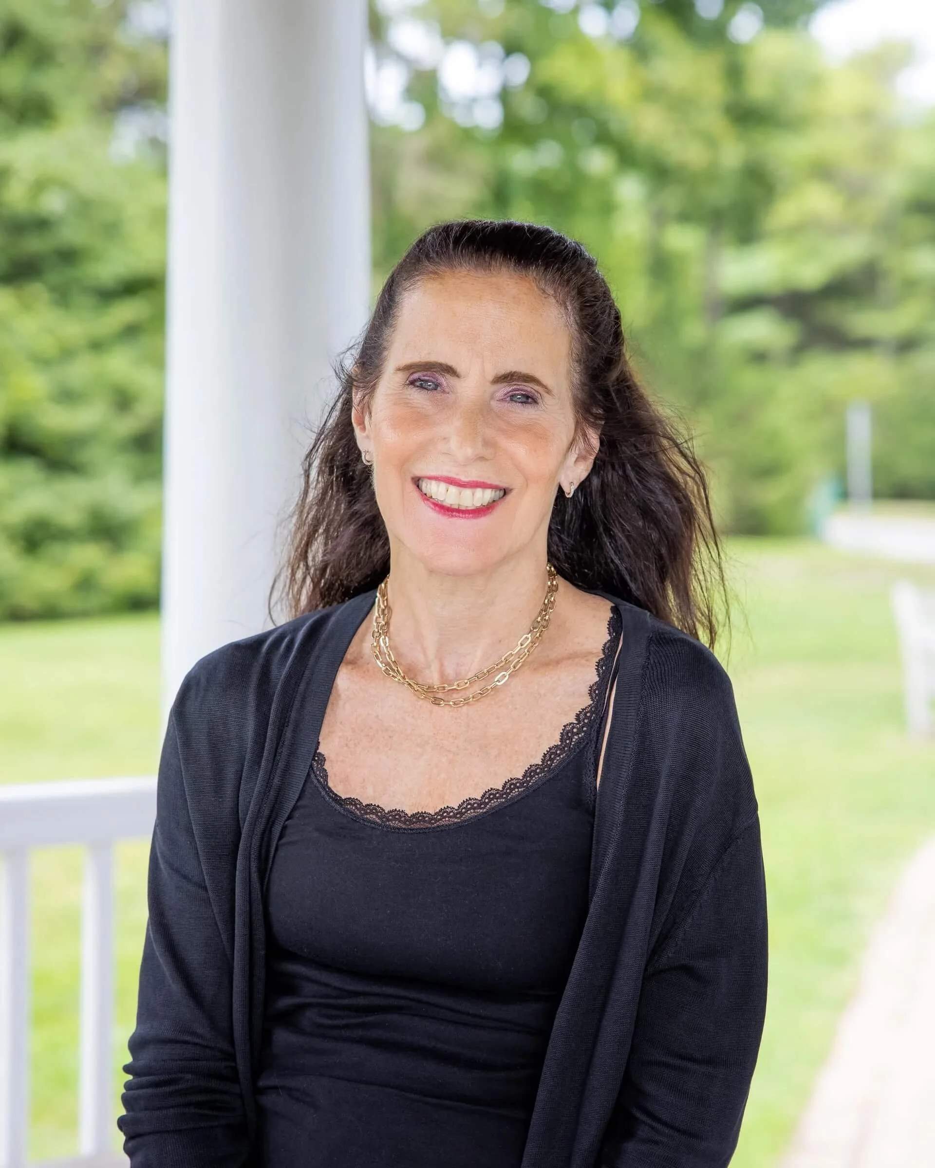 Smiling woman with long dark hair wearing a black top and a gold chain necklace, standing outdoors near a white porch railing with a green background of trees and grass.