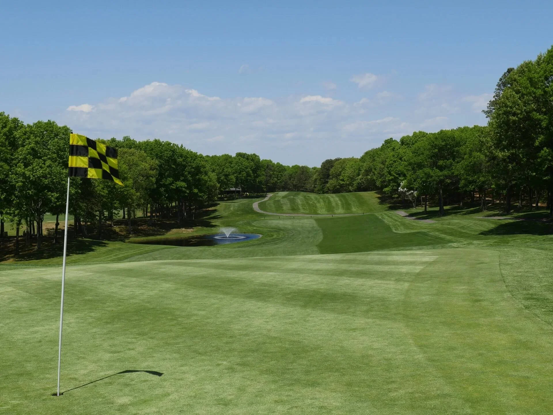 A golf course green with a yellow and black checkered flag on a pole near the hole, a water hazard with a fountain behind the green, and a tree-lined fairway under a partly cloudy blue sky.