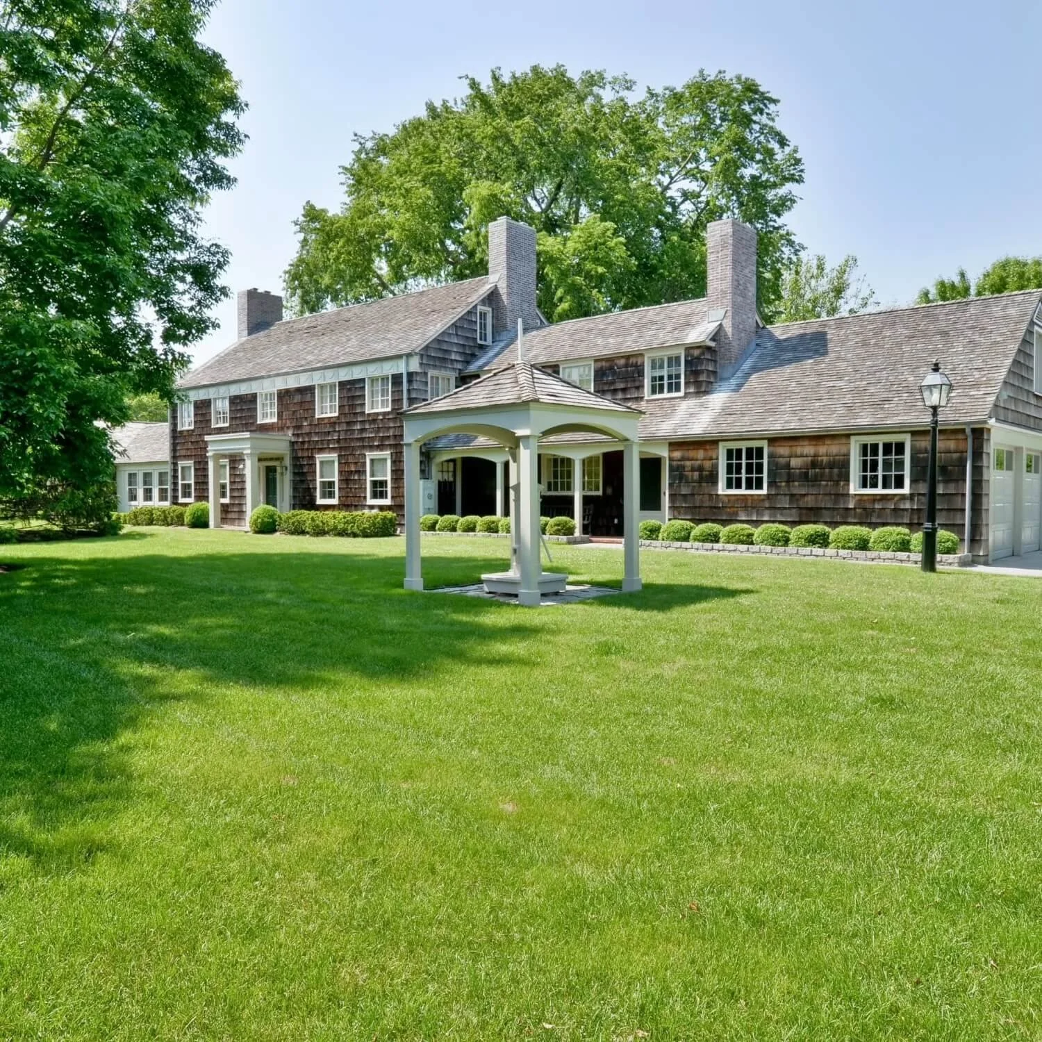 A large traditional house with dark wooden shingles, multiple chimneys, and white window frames. The house is surrounded by a well-maintained green lawn with a white wishing well at the center and a black lamp post nearby. Tall trees are in the background on a clear, sunny day.