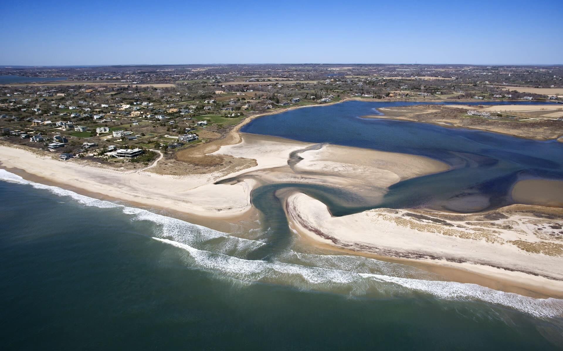 Aerial view of a coastal area with beaches, a river flowing into the ocean, and residential homes near the shoreline, under a clear blue sky.