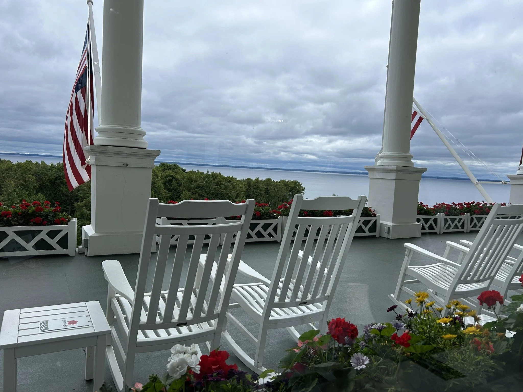 Porch with white rocking chairs, flower pots with red, yellow, and purple flowers, and American flags, overlooking a body of water under a cloudy sky.