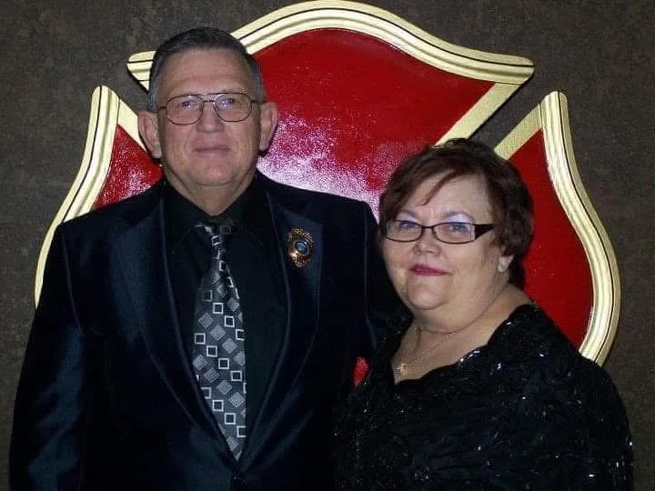 A man and woman standing together in front of a red and gold emblem or shield.