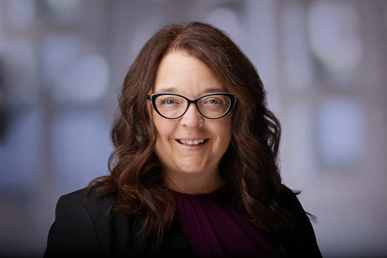 A woman with wavy brown hair, wearing glasses, smiling, dressed in a black jacket and purple top, against a blurred background.