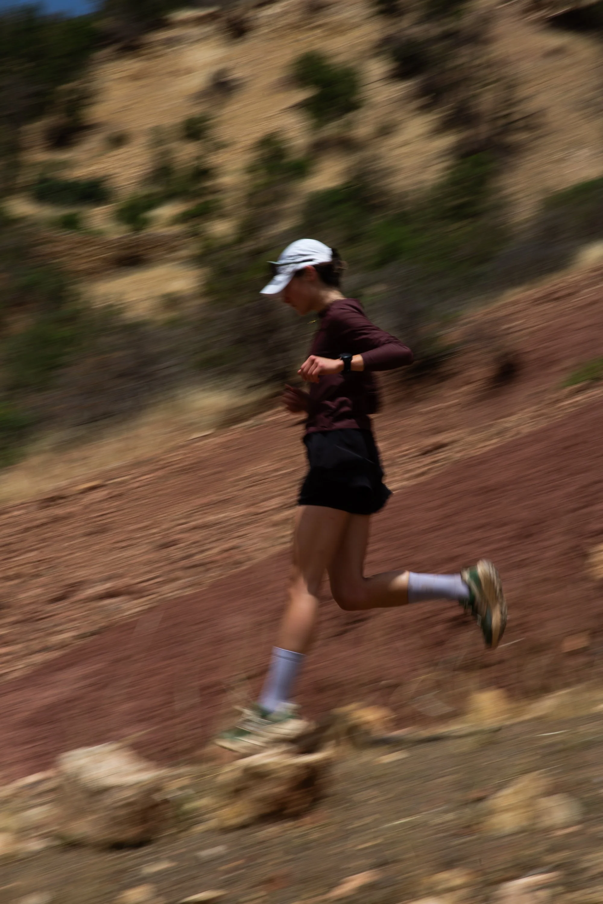 A woman running outdoors on a dirt trail along a hillside with sparse vegetation. She is wearing a white cap, a maroon long-sleeve shirt, black shorts, and running shoes, with a watch on her left wrist.