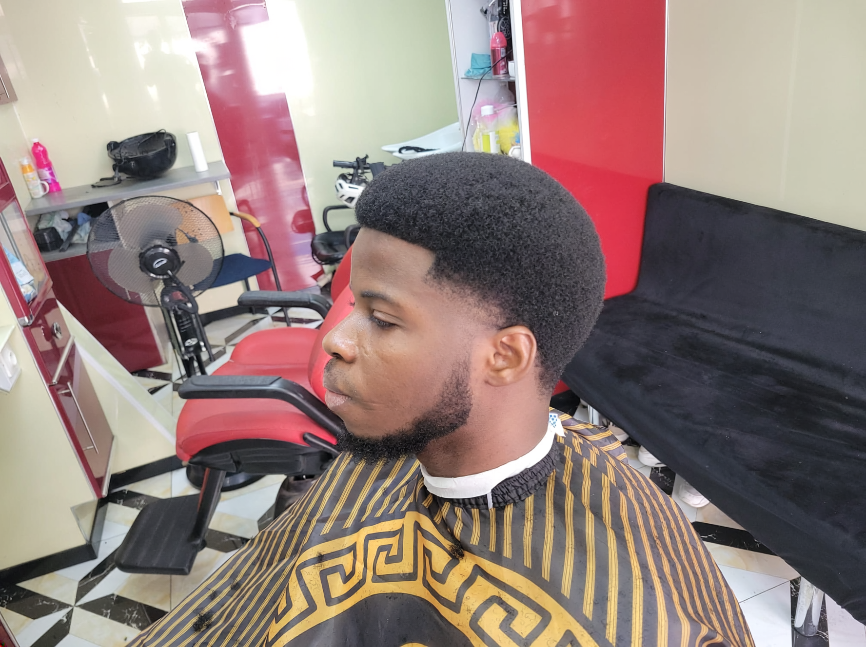 Jeune homme avec une coupe afro nette dans un salon de coiffure, portant une cape de coiffure à motifs dorés.
