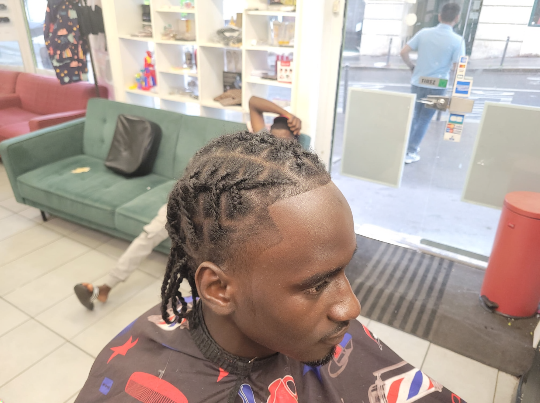 Un homme avec des tresses à l'avant dans un salon de coiffure, assis sur une chaise, avec une autre personne allongée à l'arrière, portant un casque audio, devant une vitrine avec des produits et une porte en verre.