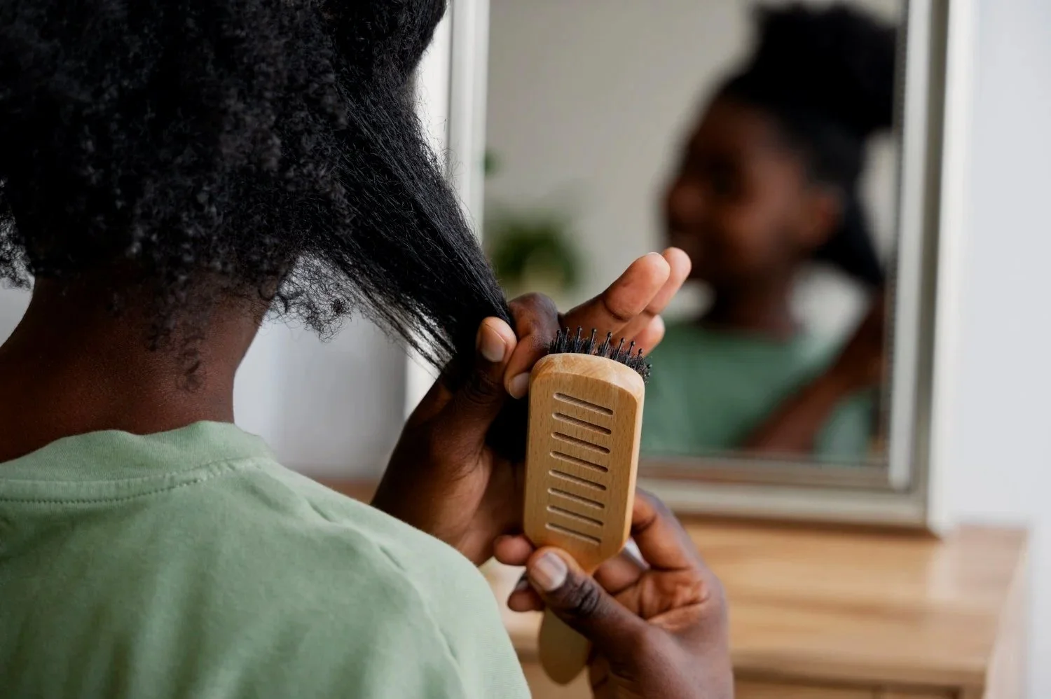 Une femme se brosse les cheveux avec une brosse en bois devant un miroir, reflet de son visage flou.