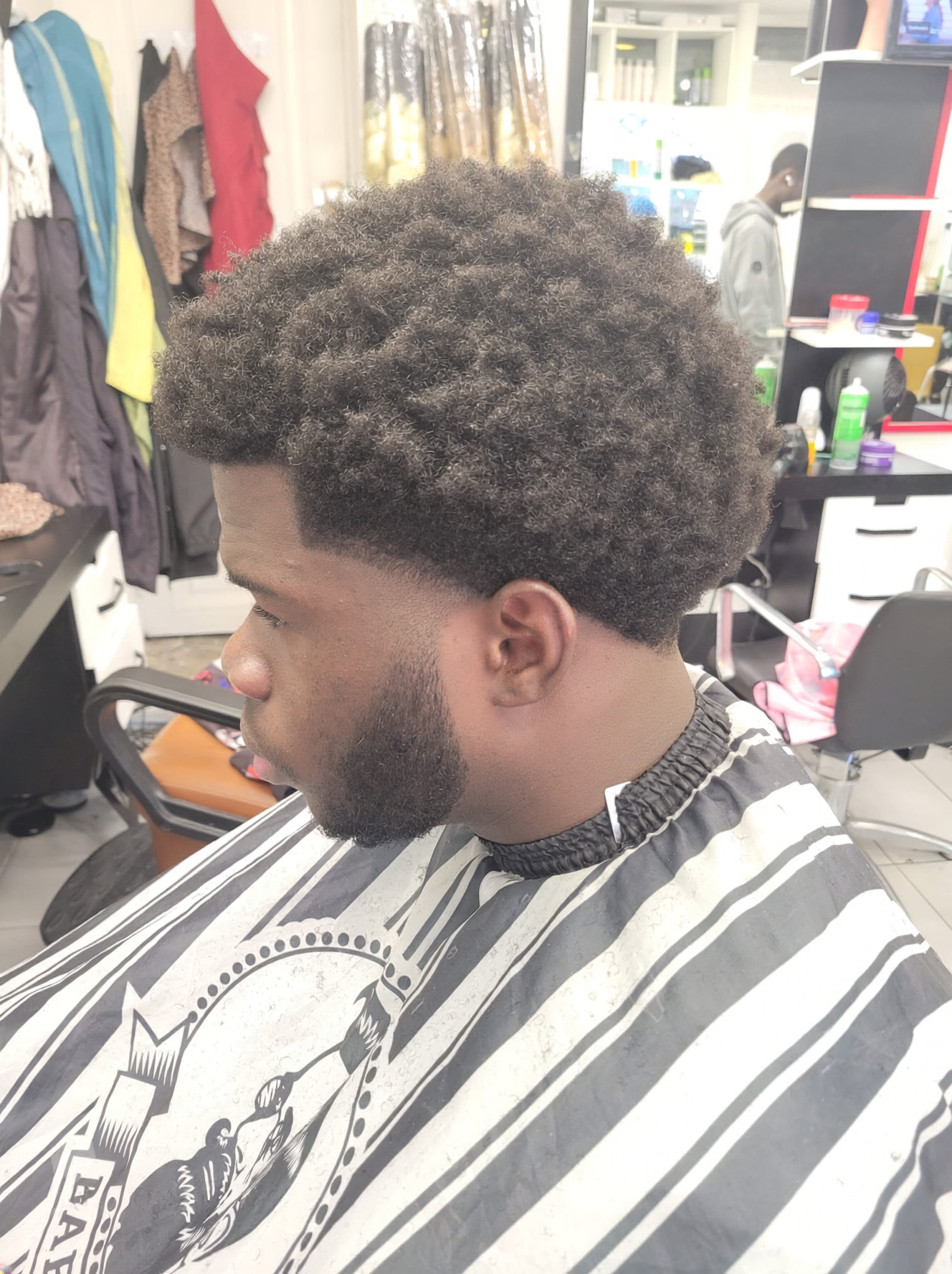 Jeune homme avec une coiffure en afro et barbe, assis dans un salon de coiffure avec un peignoir à rayures noires et blanches.