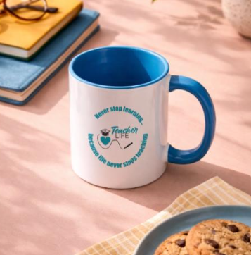 A white ceramic mug with blue handle and interior, featuring a motivational message and design, placed on a desk with a plate of cookies and notebooks.
