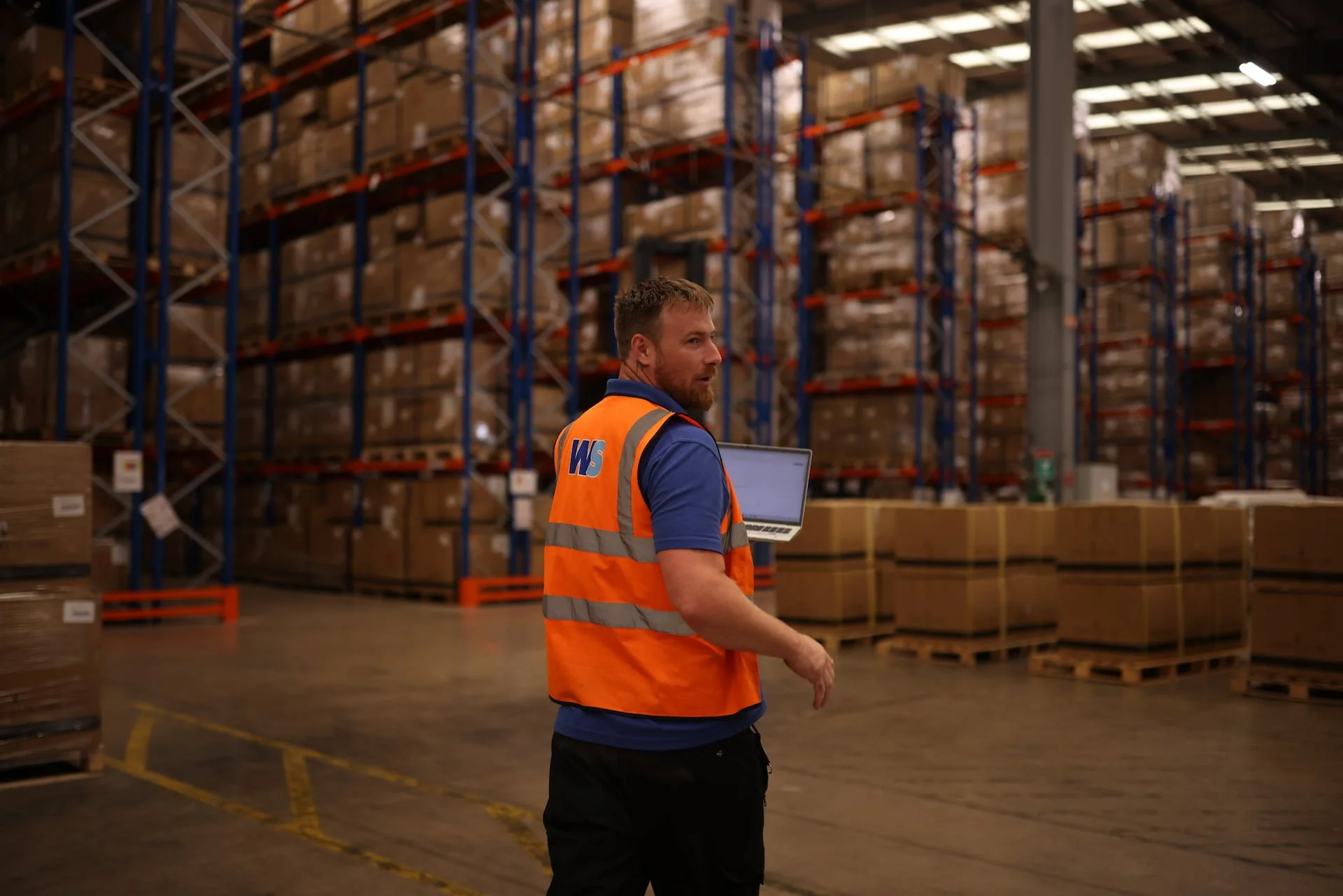 A warehouse worker wearing an orange safety vest and a blue shirt holds a laptop while inspecting storage shelves filled with boxes.