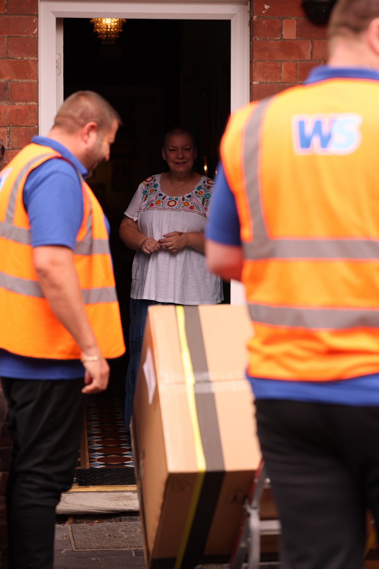 A woman stands inside her doorway smiling as two delivery workers in orange safety vests with the letters 'WS' on the back deliver a package outside in the evening.