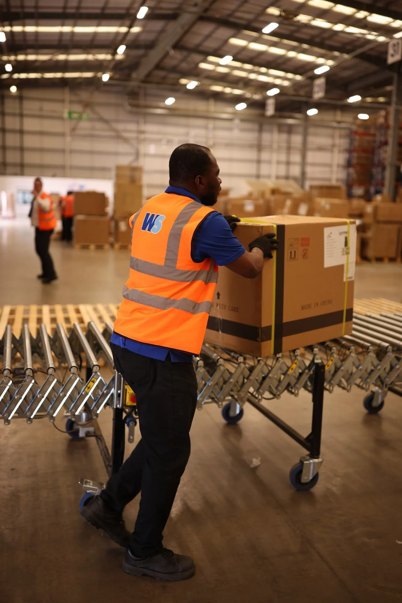 A warehouse worker wearing an orange safety vest with blue and white logos, black gloves, and black pants pushes a box on a roller conveyor in a large industrial warehouse with shelves and additional workers in the background.