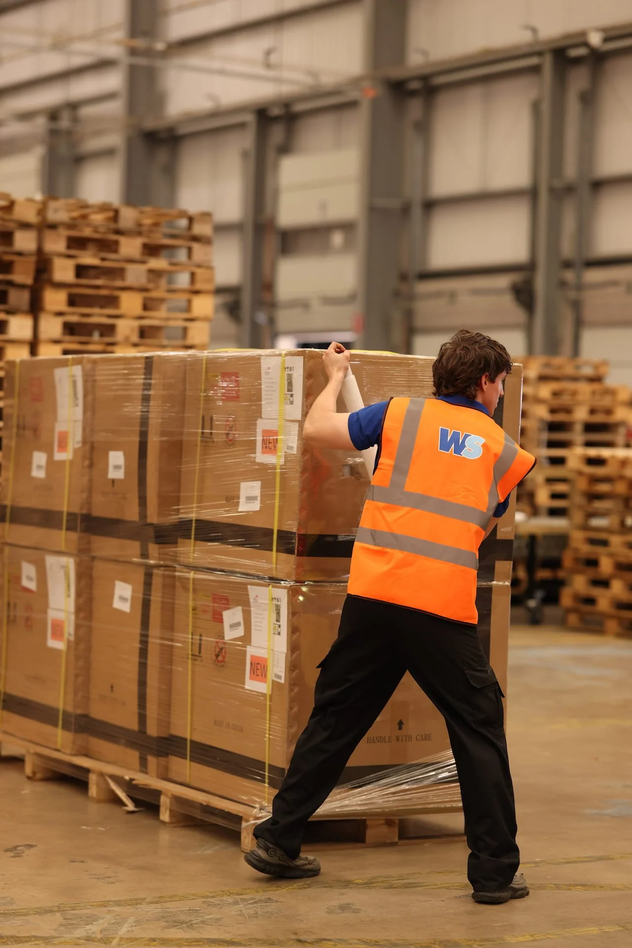 A warehouse worker in an orange safety vest and black pants is pulling a large wrapped pallet of boxes on a wooden pallet.