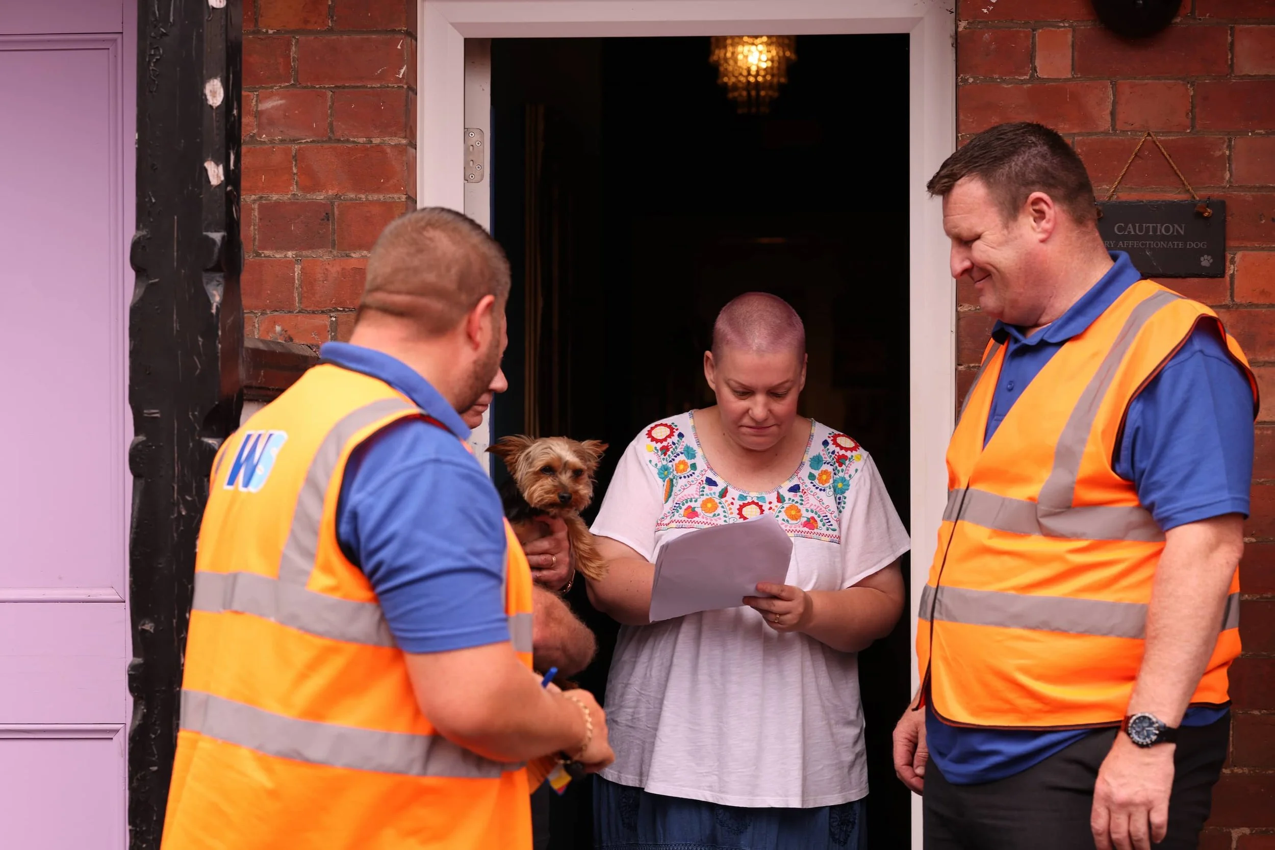 A woman with a shaved head holding a small dog and looking at a piece of paper, standing at her door as two men in orange safety vests and blue shirts talk to her.
