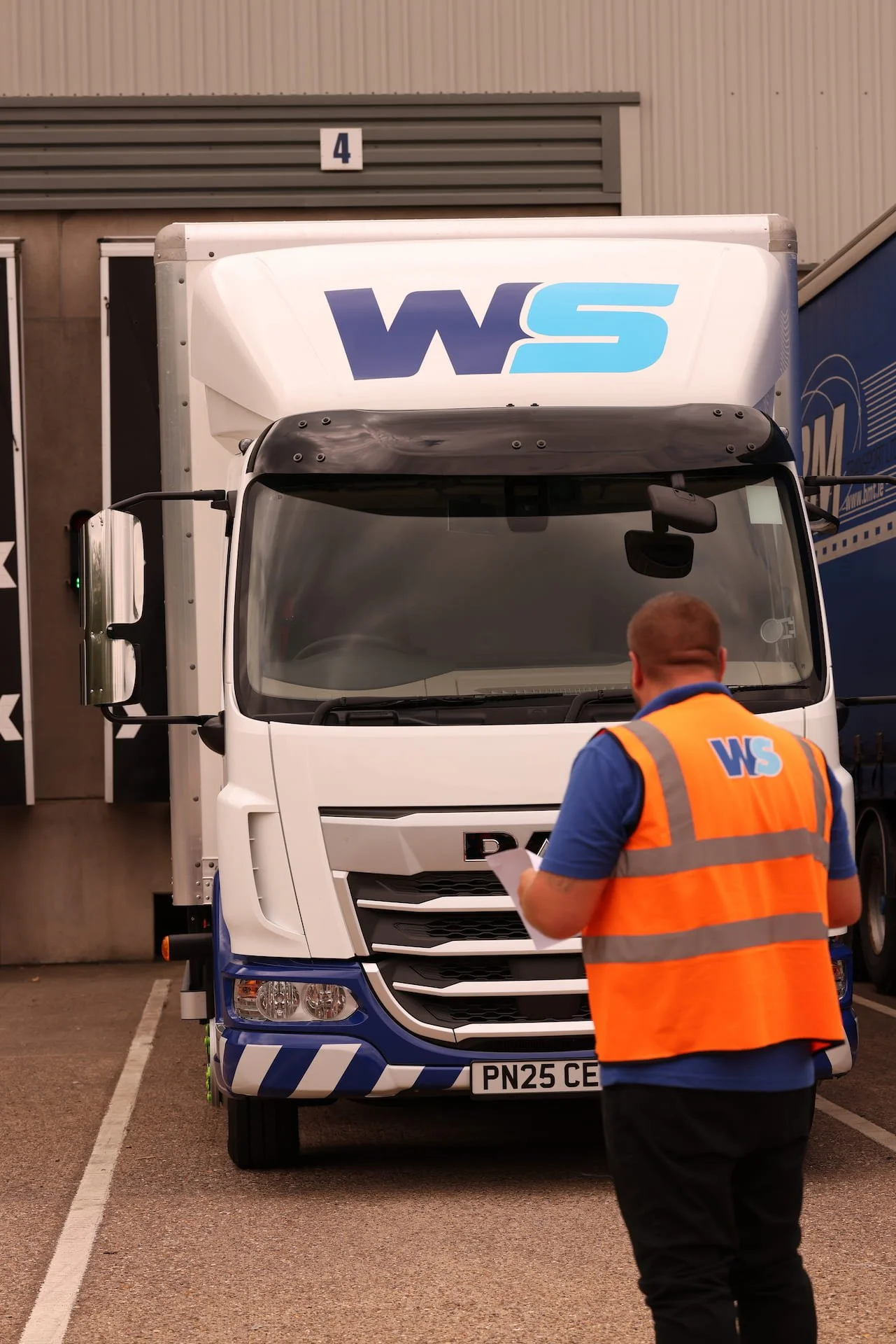 A man in an orange vest with WS logo looking at his phone in front of a delivery truck with WS logo.