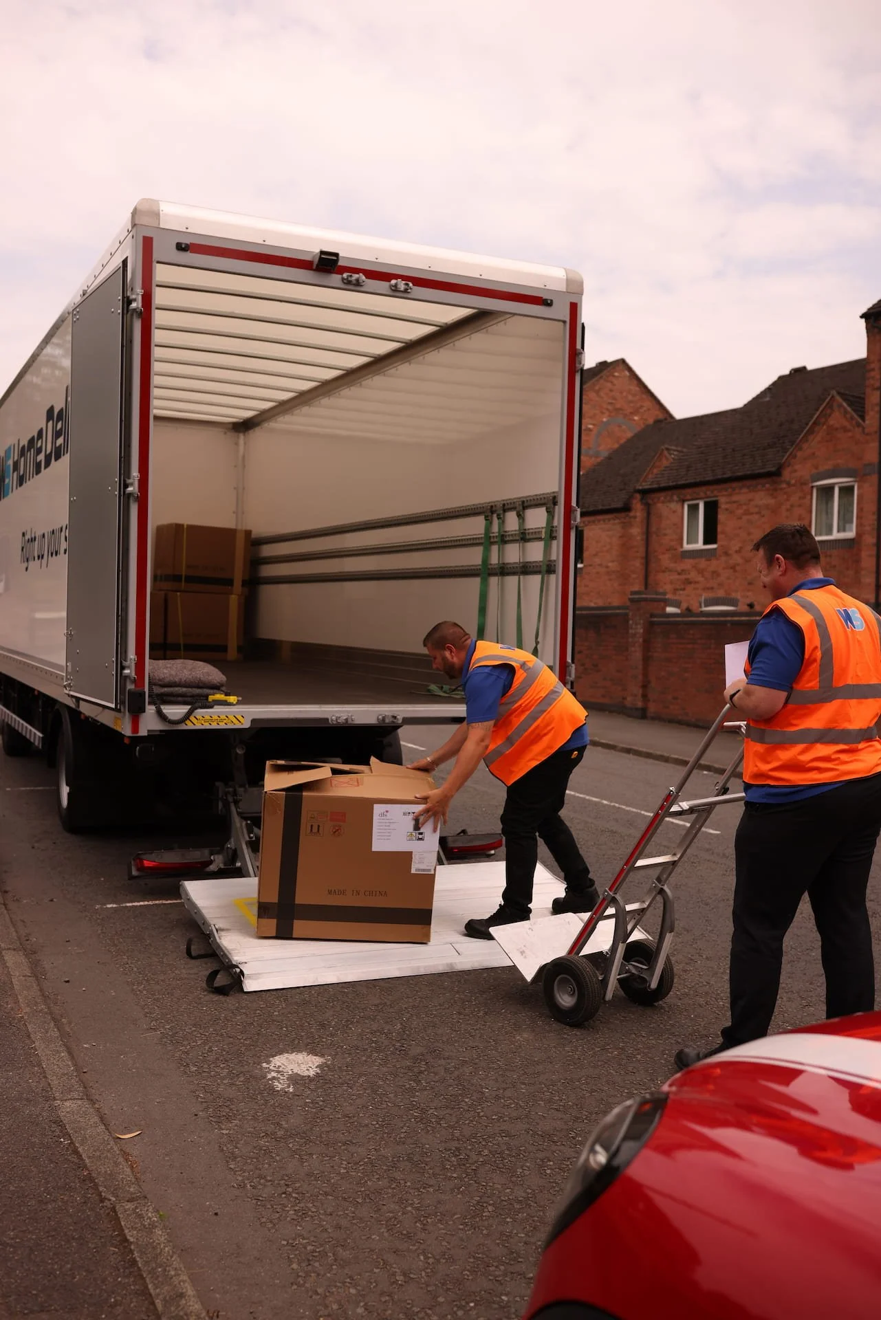 Two movers loading a large cardboard box into a moving truck parked on the street.