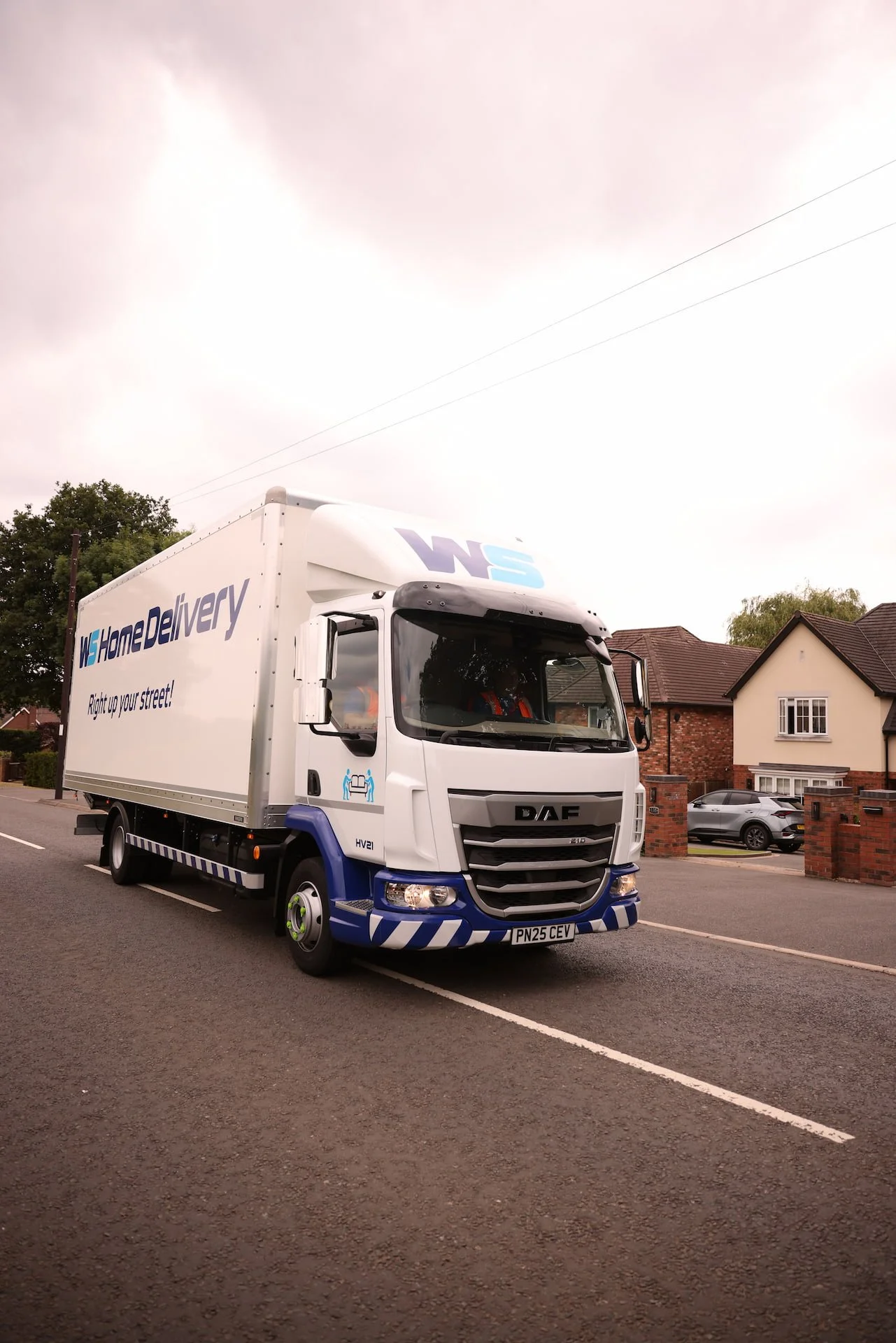 A white delivery truck with blue accents and the logo 'WHS Home Delivery' is parked on a residential street, with houses and parked cars in the background under an overcast sky.