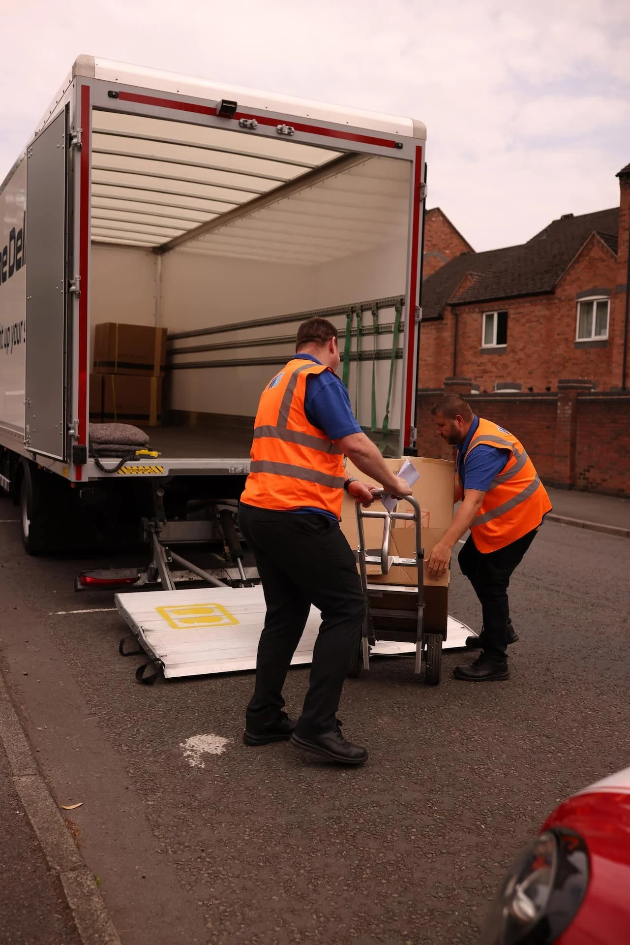Two men wearing orange safety vests loading boxes into a delivery truck on a street with brick houses in the background.