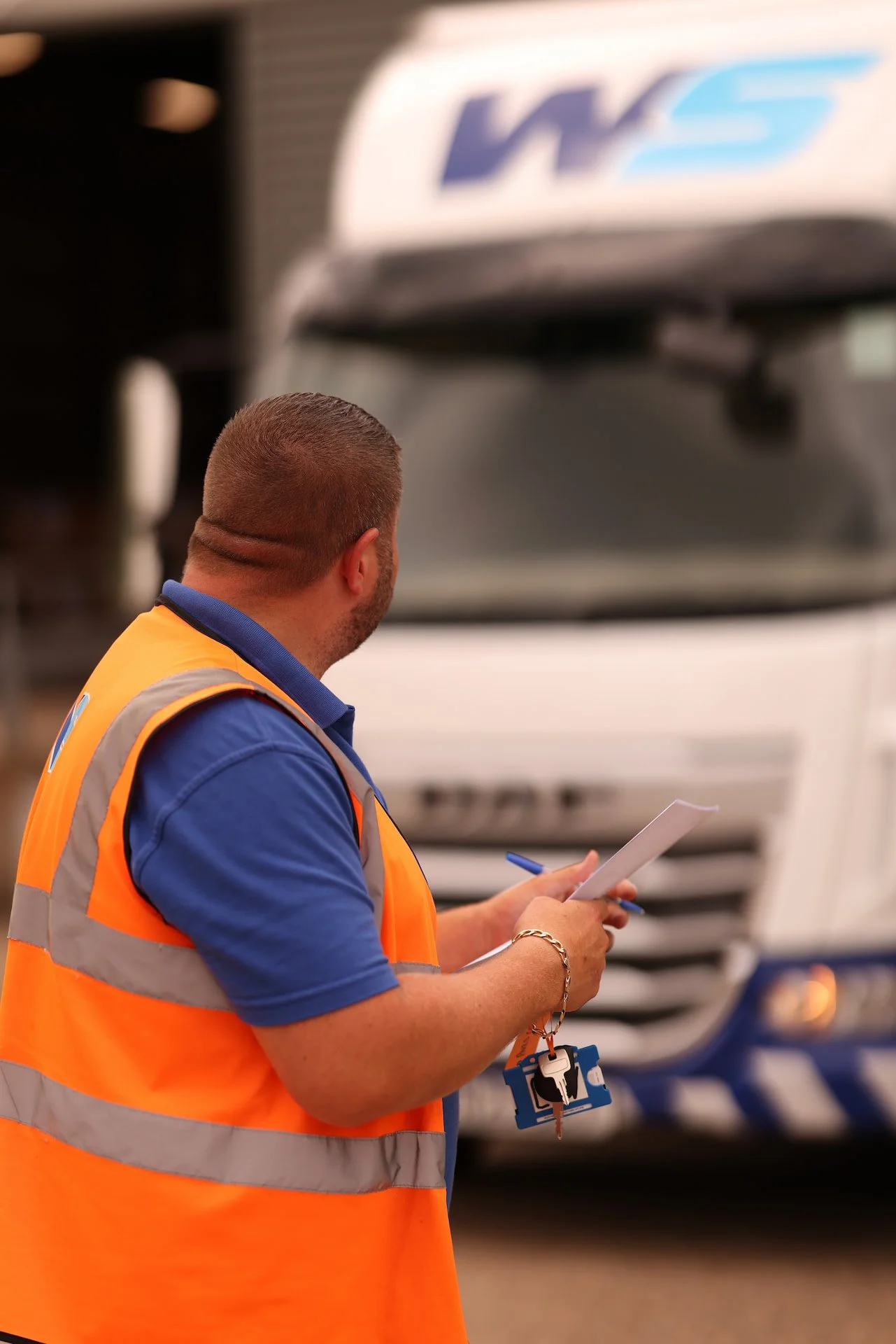 A man with a beard, wearing a blue polo shirt and orange safety vest, is holding a notepad and a pen, standing in front of a large white semi-truck, while looking away from the camera.