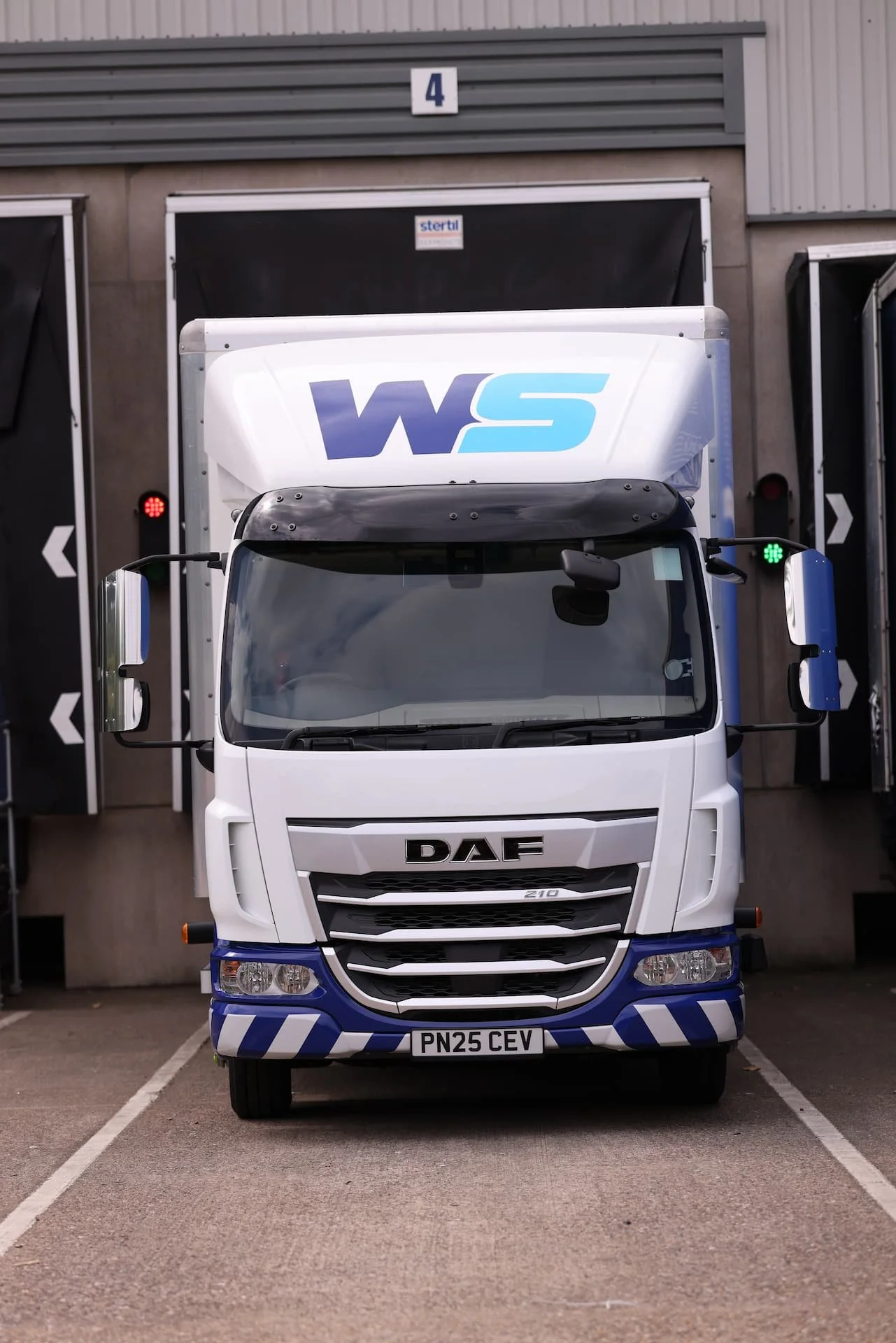 Front view of a DAF truck with a white cab and blue accents, parked in front of a loading dock with number 4, showing green and red lights.