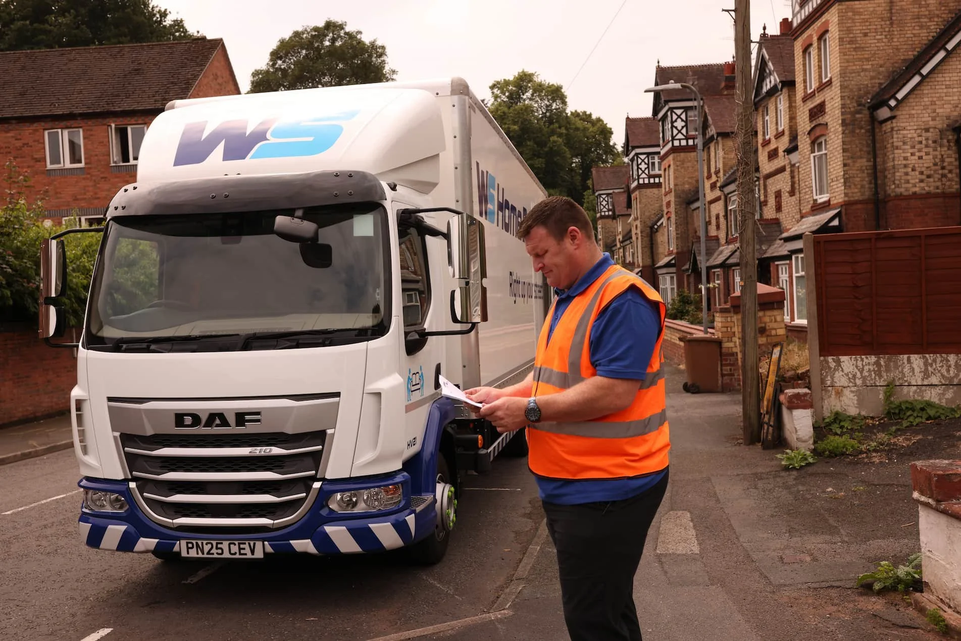 A delivery man in an orange safety vest standing beside a white delivery truck, looking at a clipboard in a residential neighborhood.