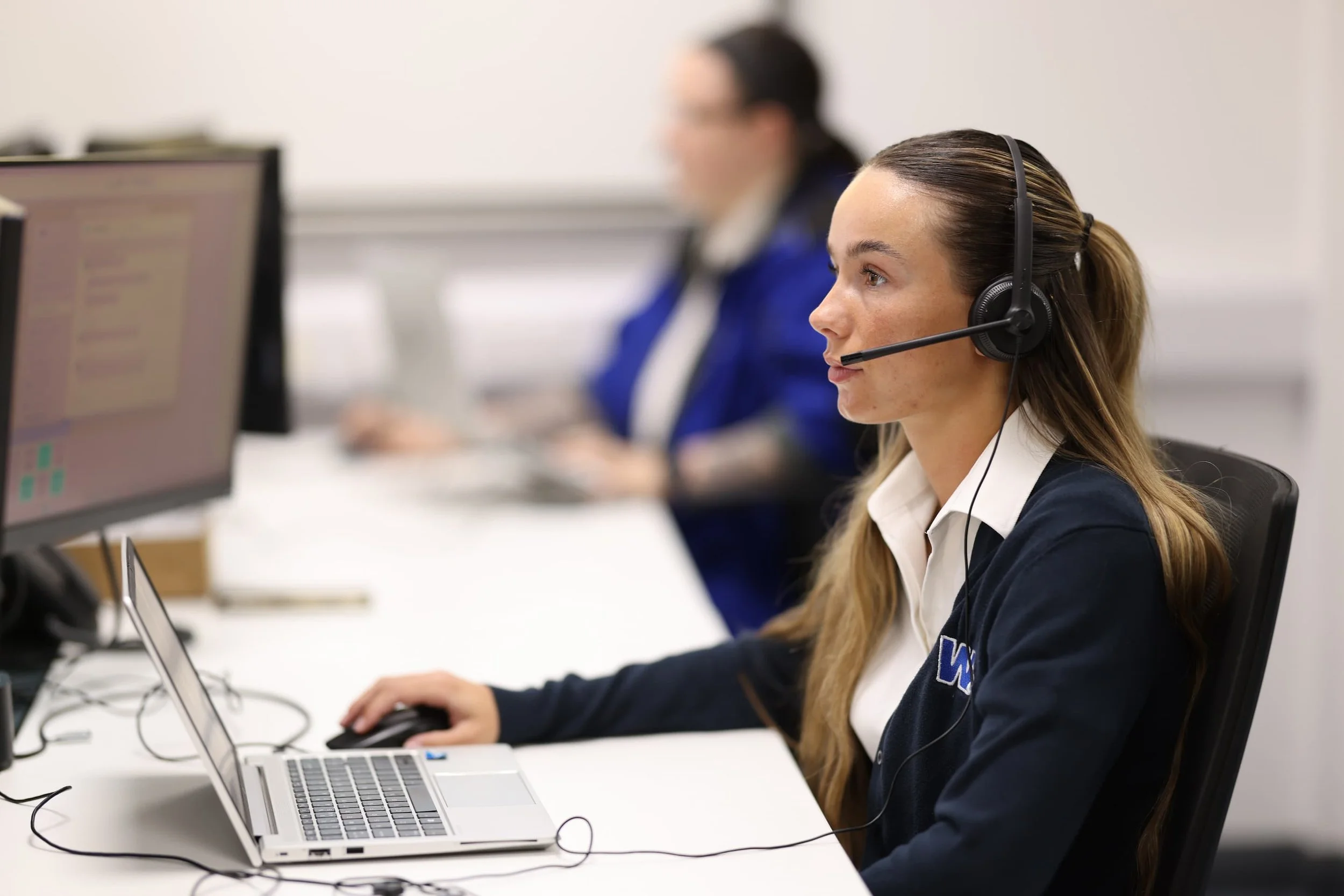 A woman wearing a headset and a navy blue cardigan working at a computer in an office.