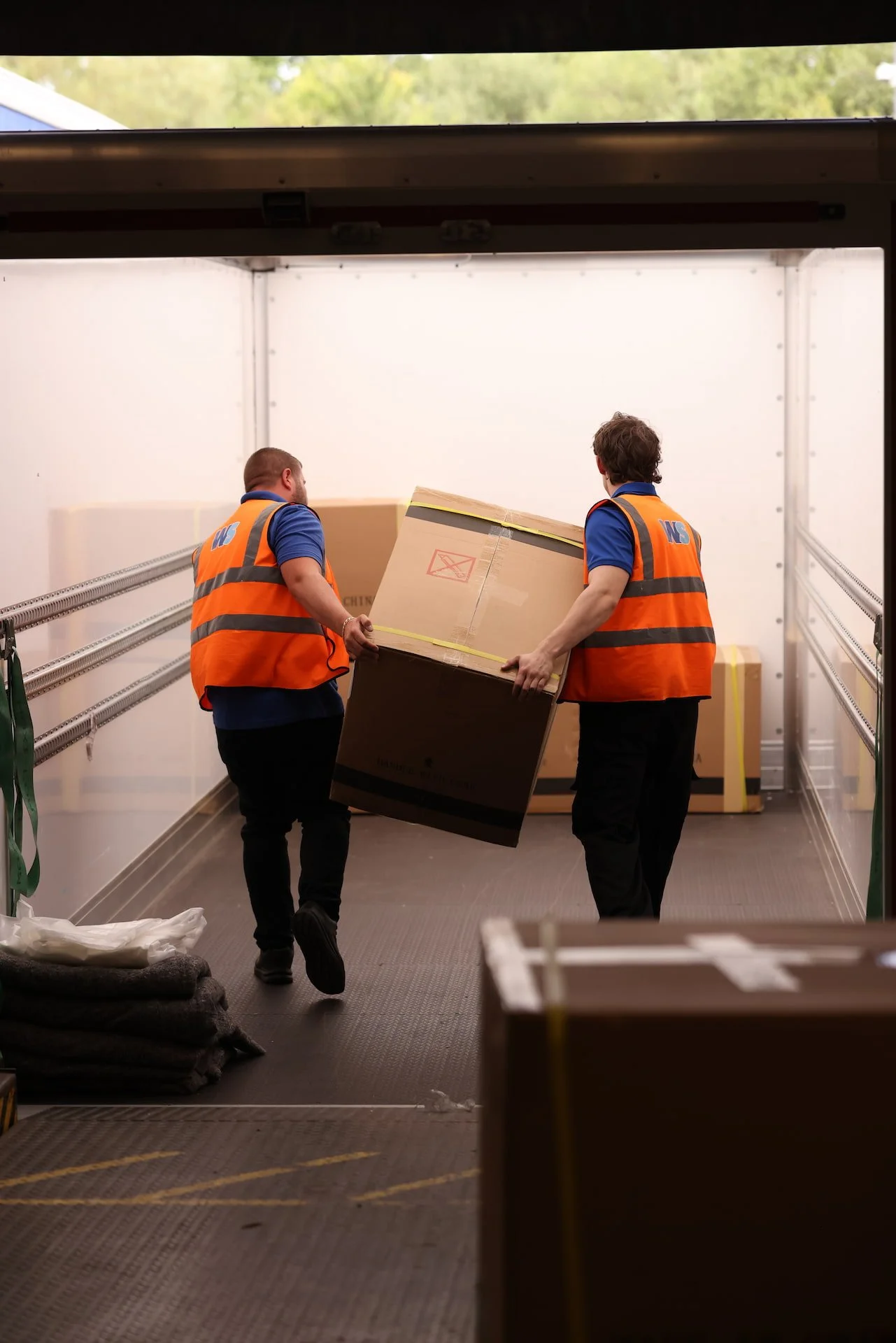 Two workers in orange safety vests load a large cardboard box into a delivery truck.