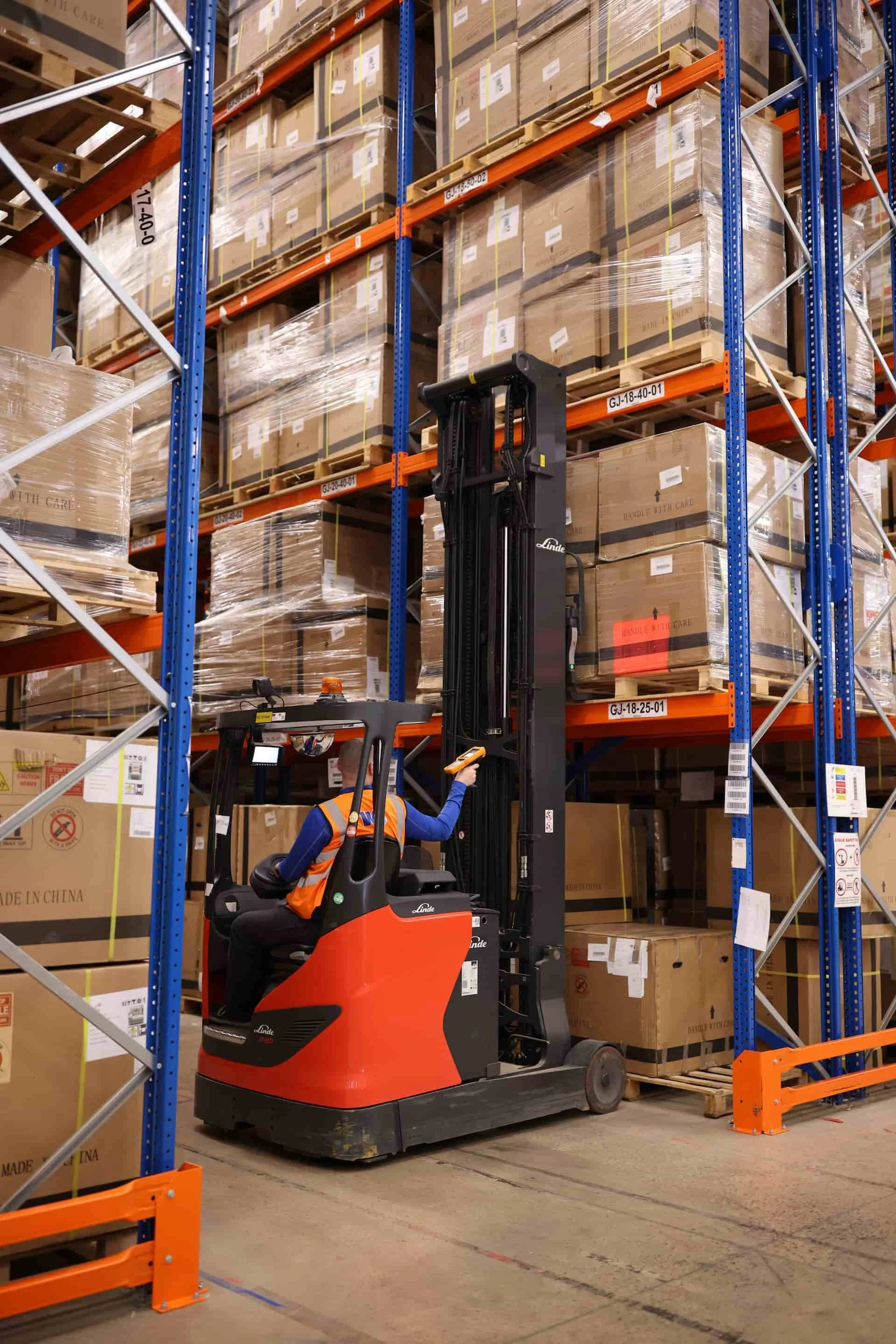 A worker operating a forklift in a large warehouse filled with stacked cardboard boxes on metal shelves. The forklift is red and black, and the worker is wearing a blue uniform and an orange safety vest.