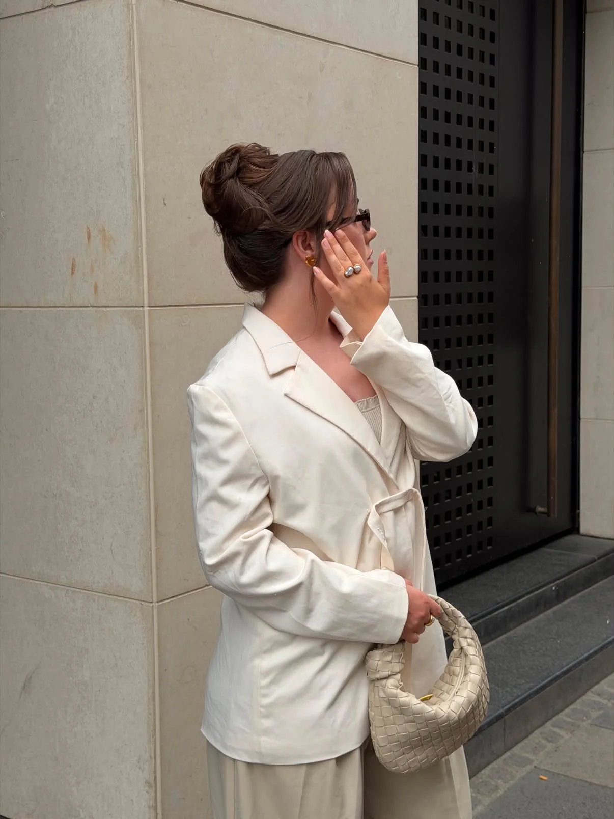 A woman with brown hair styled in an updo, wearing glasses and a cream-colored blazer, standing against a stone wall near an elevator, holding a beige woven handbag.
