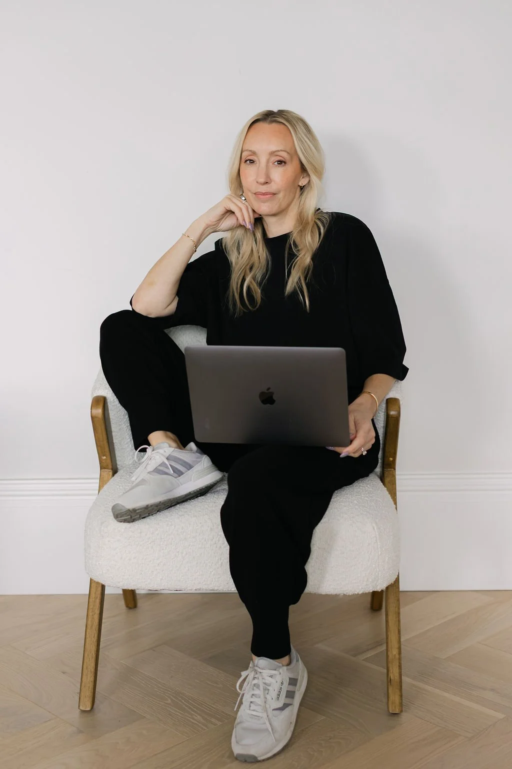 A woman with long blonde hair sat on a chair with one foot up and a laptop on her legs
