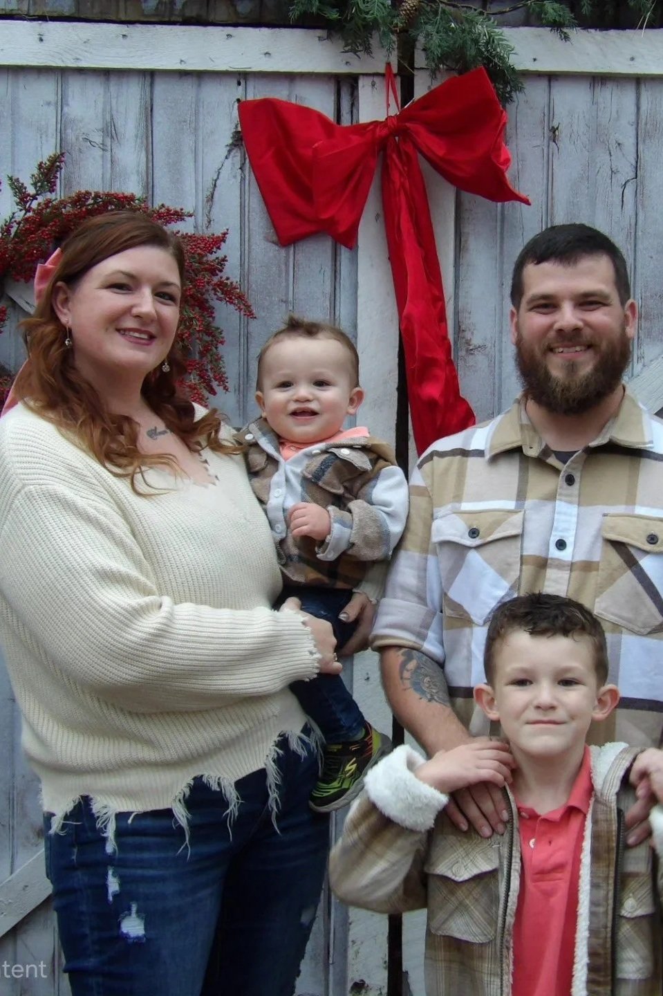 A family of four standing outdoors in front of a wooden fence decorated with a large red bow and a wreath, celebrating a holiday.
