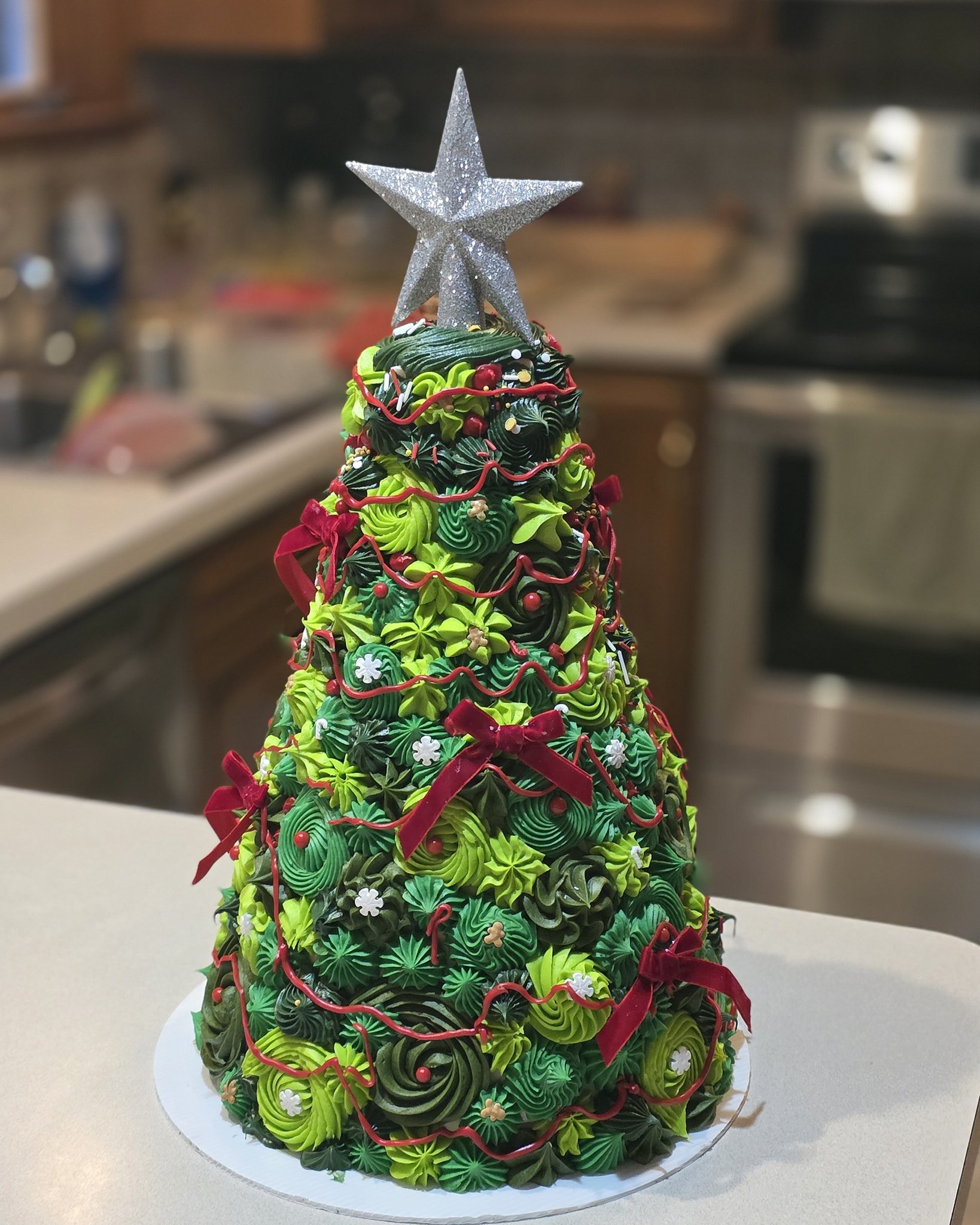A Christmas tree decorated with green icing in various swirls and patterns, red bows, small white and red ornaments, and topped with a silver glittery star.