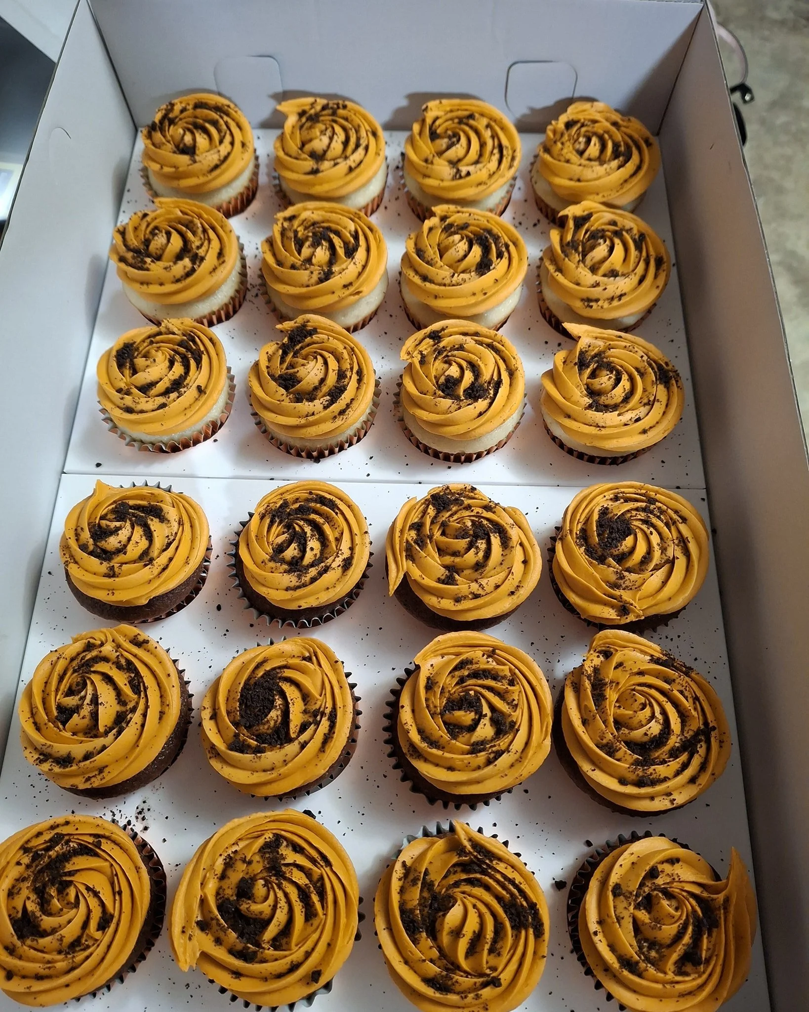 Box of 24 cupcakes with yellow frosting and chocolate cookie crumbles on top.
