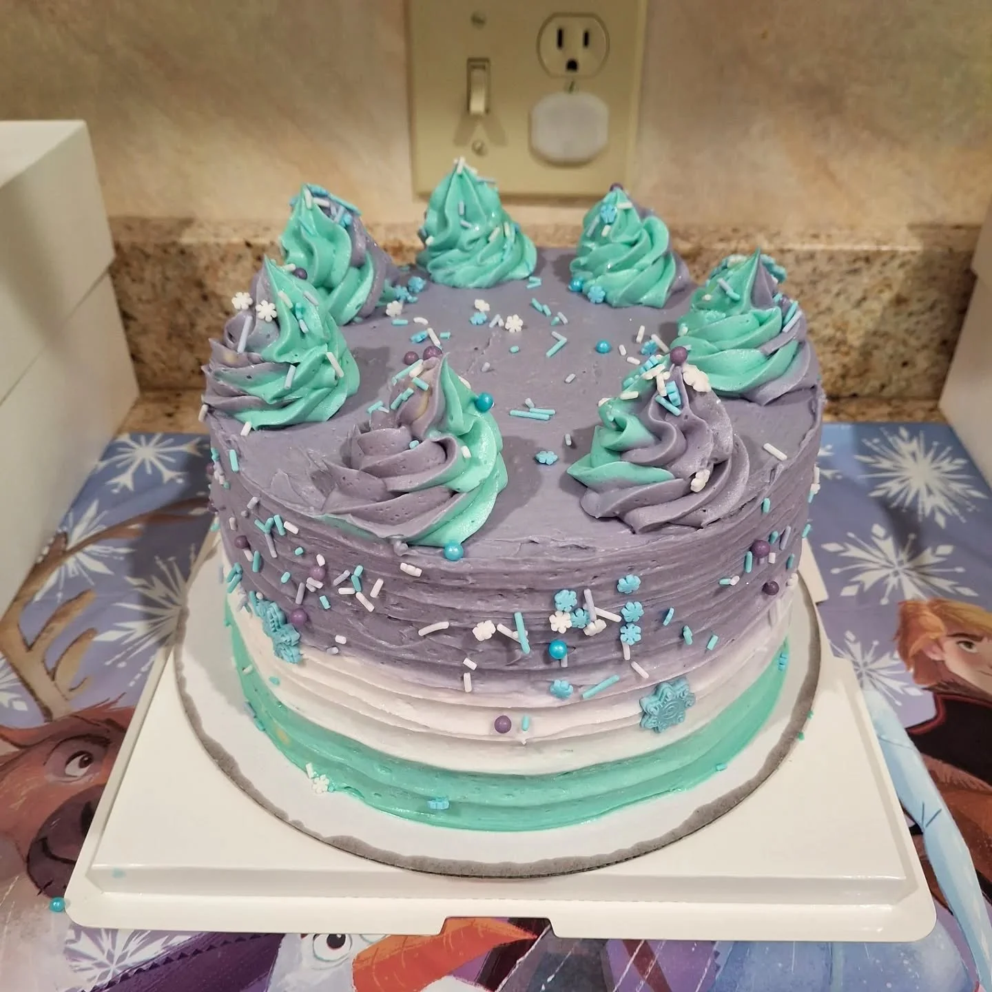 A birthday cake decorated in shades of purple and turquoise with swirl frosting, sprinkles, and snowflake-shaped decorations, set on a white square cake board.