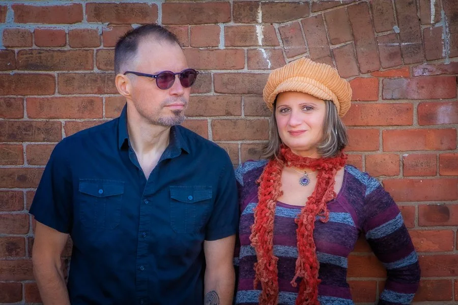 A man and woman standing against a brick wall. The man is wearing sunglasses and a dark blue short-sleeved shirt. The woman is wearing a beige beret, a colorful striped top, a red scarf, and jewelry.