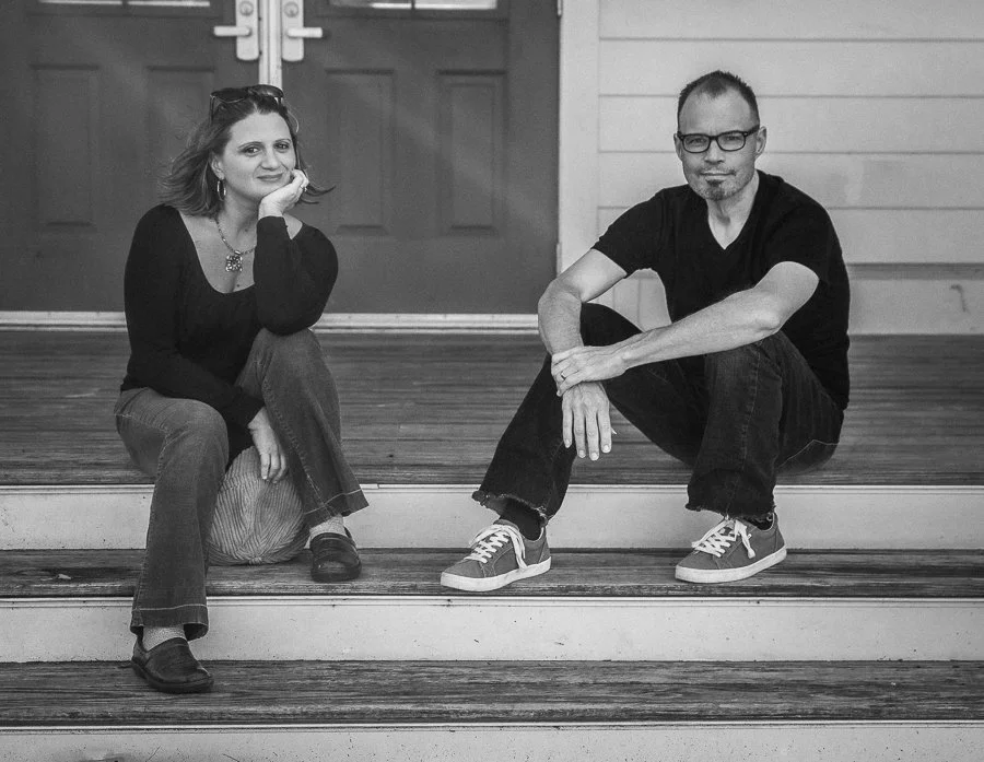 A woman and a man sitting on wooden steps outside a building, both casually dressed, with the woman resting her chin on her hand and smiling, and the man sitting with one arm around his knee and wearing glasses.