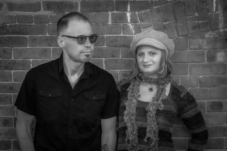 A black and white photo of a man and woman standing in front of a brick wall. The man is wearing sunglasses and a black shirt, while the woman is wearing a beret, a striped shirt, and a scarf.