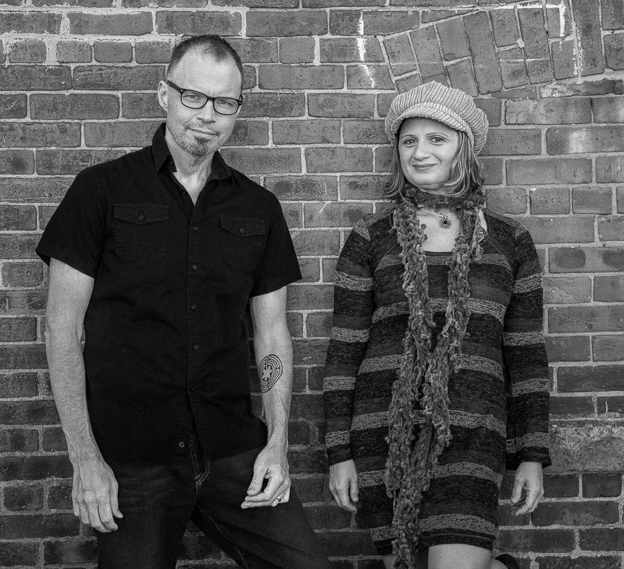 A man and woman standing against a brick wall, both smiling. The man is wearing glasses and a black short-sleeved shirt, while the woman is wearing a beret, a striped dress, and a long scarf.